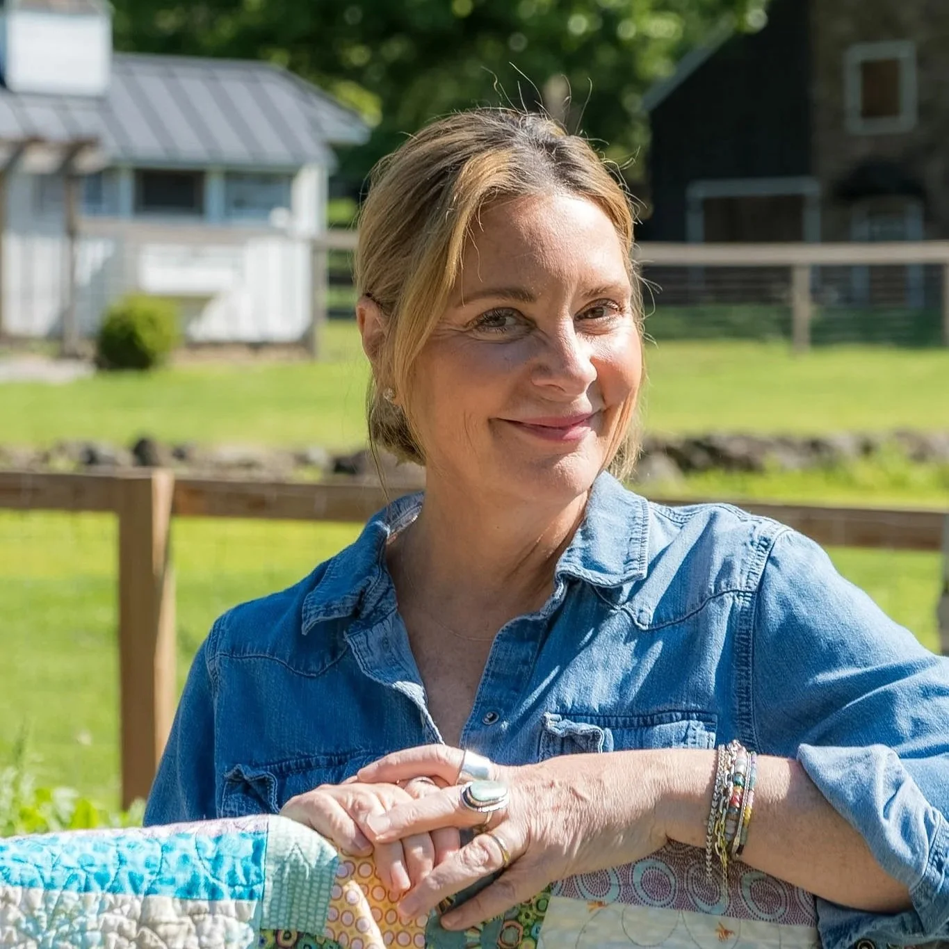 A woman with short blonde hair smiling outdoors, wearing a blue denim shirt, with a wooden fence and houses in the background.