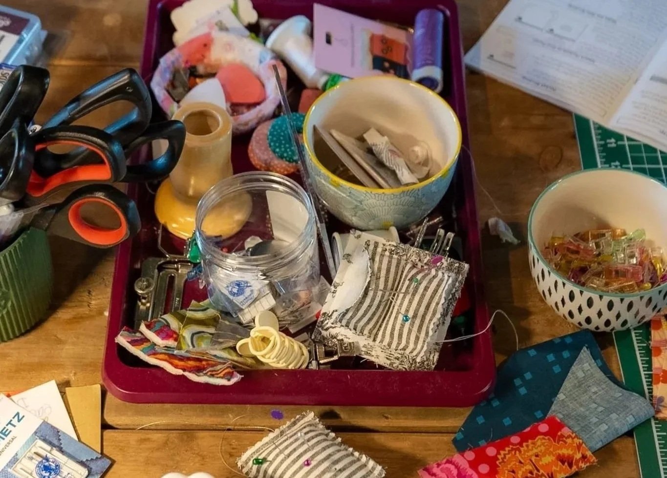 A cluttered sewing or craft workspace with various sewing supplies including scissors, fabric scraps, bowls with sewing notions, thread, and other crafting materials on a wooden table.