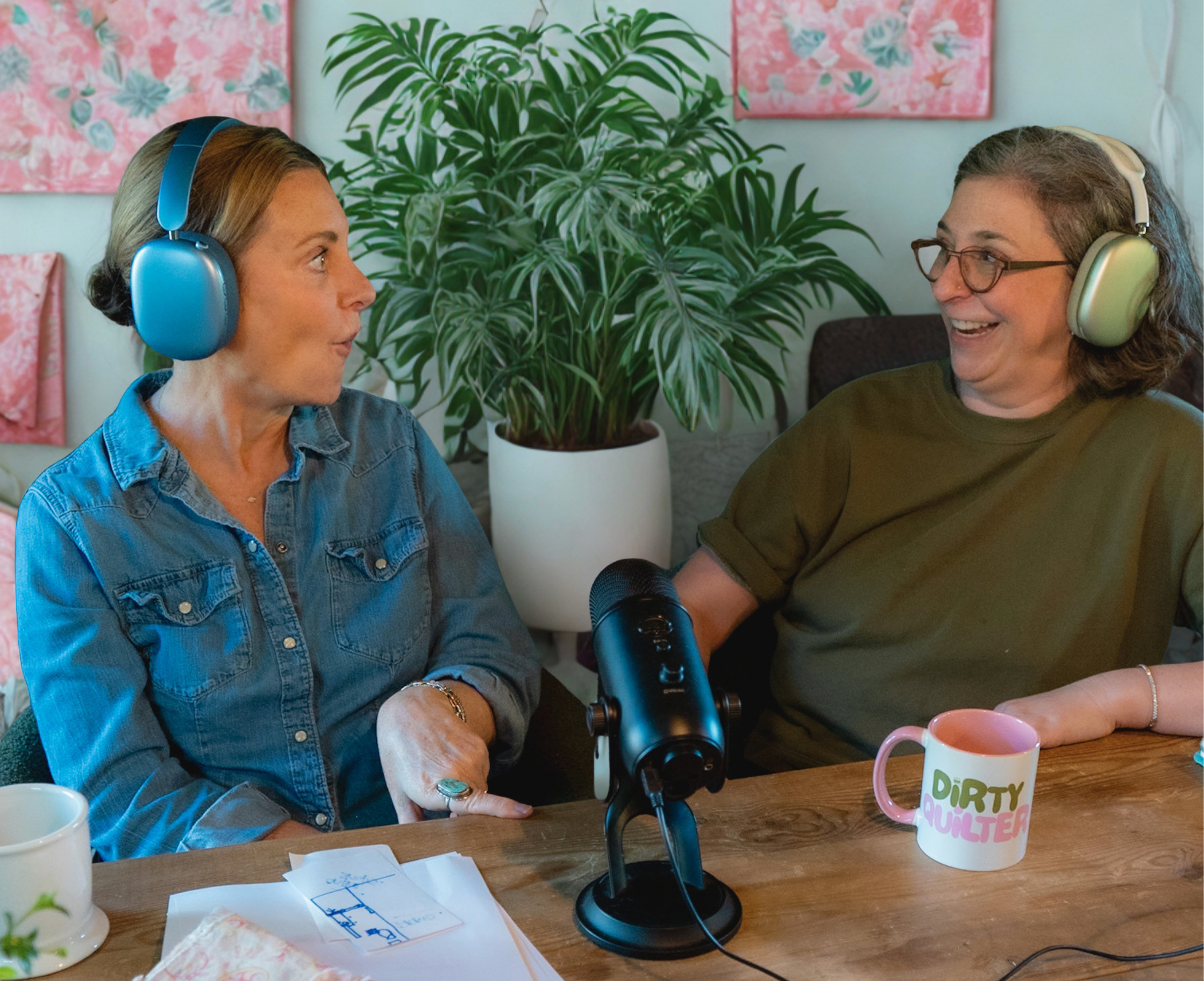 Two women sitting at a table with a large green plant in the background, wearing headphones, and looking at each other. There are papers and mugs on the table, one mug has colorful text that says "Dirty Quilter."