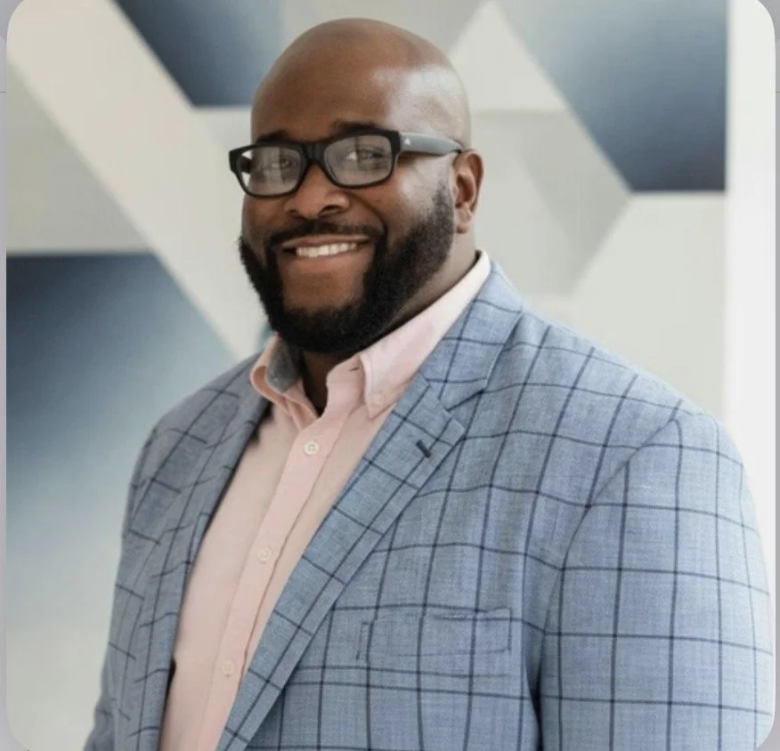 A smiling Black man with glasses, a beard, wearing a light pink shirt and a blue plaid blazer standing indoors.