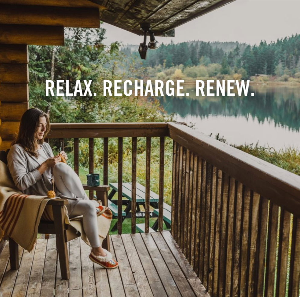 A woman sitting on a wooden porch with a railing, overlooking a lake surrounded by trees, reading a book, with a blanket and slippers, with the text 'Relax. Recharge. Renew.'