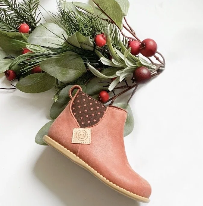A pink children's shoe with a small loop at the back and a beige sole, resting on a white surface near a holiday-themed greenery arrangement with red berries and green leaves.