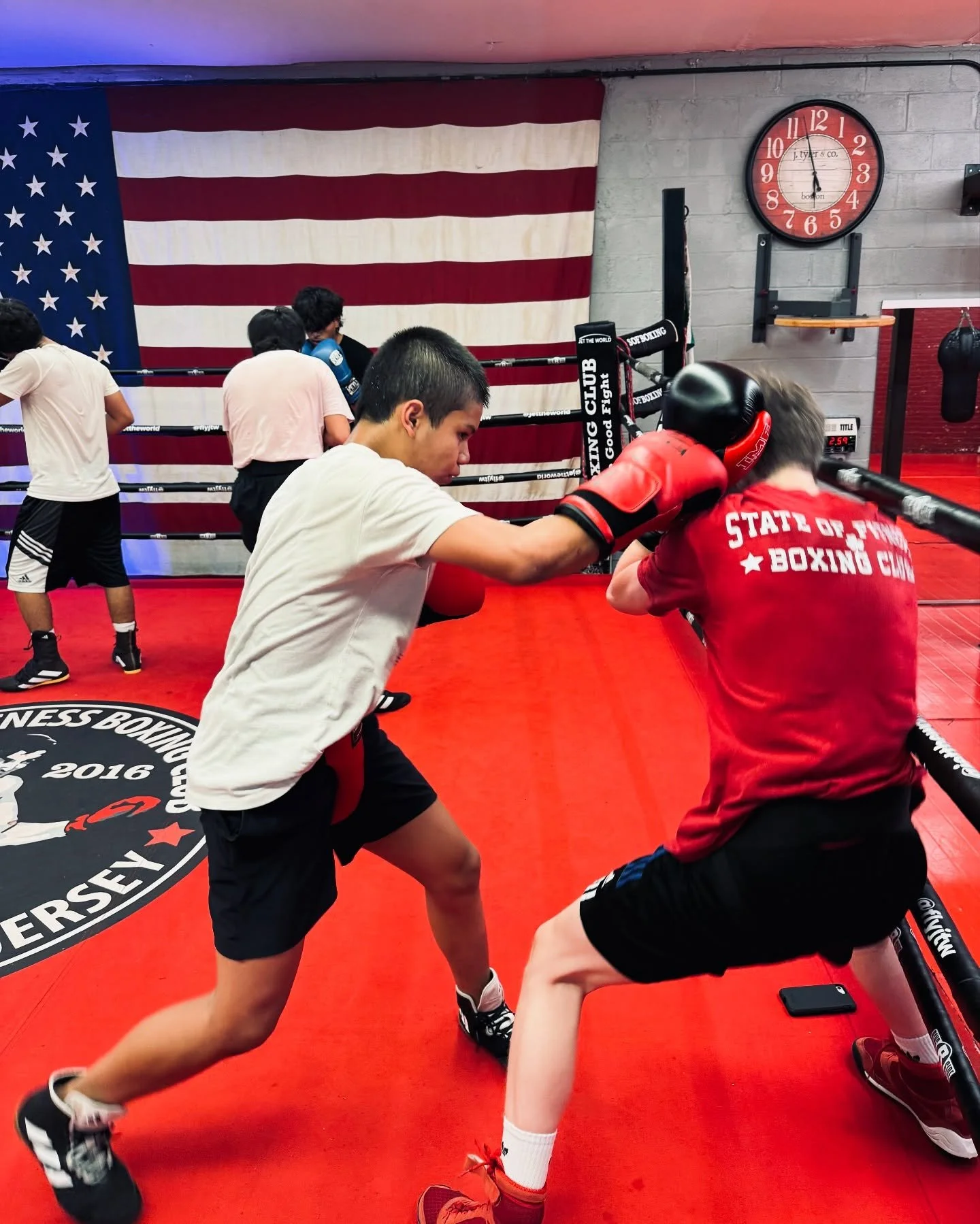 Two young men practicing boxing in a gym with American flags on the wall and a red ring in the background. One man is wearing a white t-shirt and black shorts, punching the other man wearing a red shirt and black shorts, who is holding boxing pads.