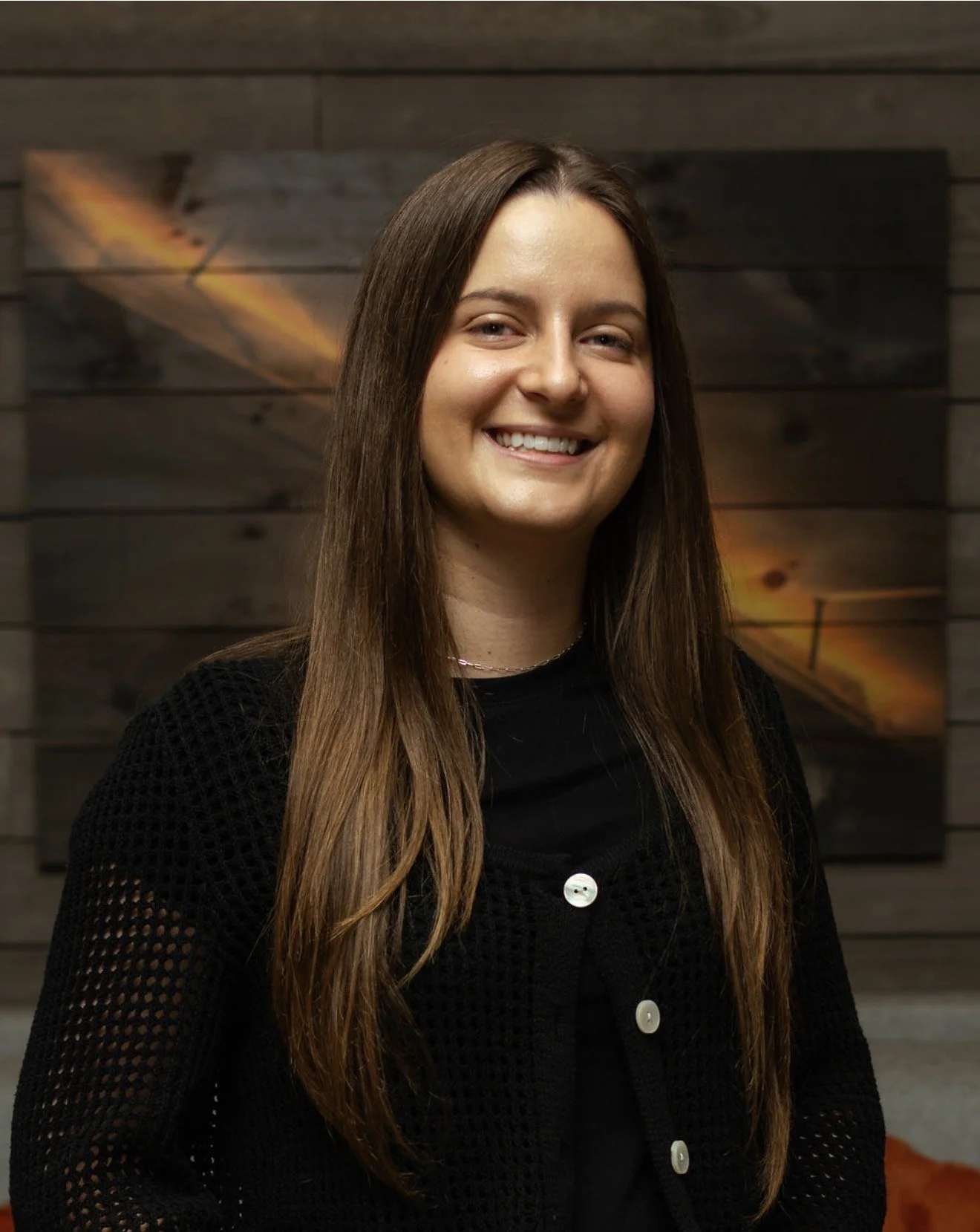 A young woman with long brown hair smiling, wearing a black knitted sweater with white buttons, standing in front of a wooden wall with a colorful abstract painting.