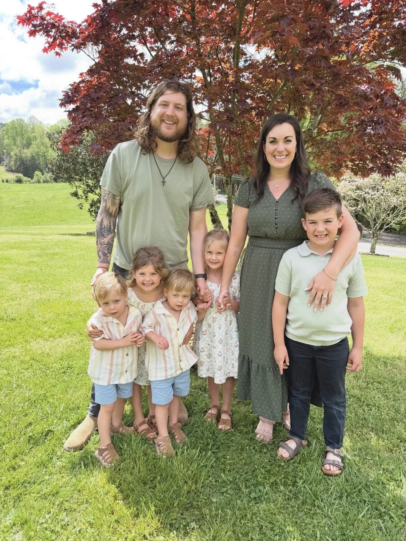 A family of seven standing outdoors on grass with trees and a blue sky in the background. Two adults, a man with long hair and a beard, and a woman with dark hair, are surrounded by five children. The children are smiling, and some are holding hands.