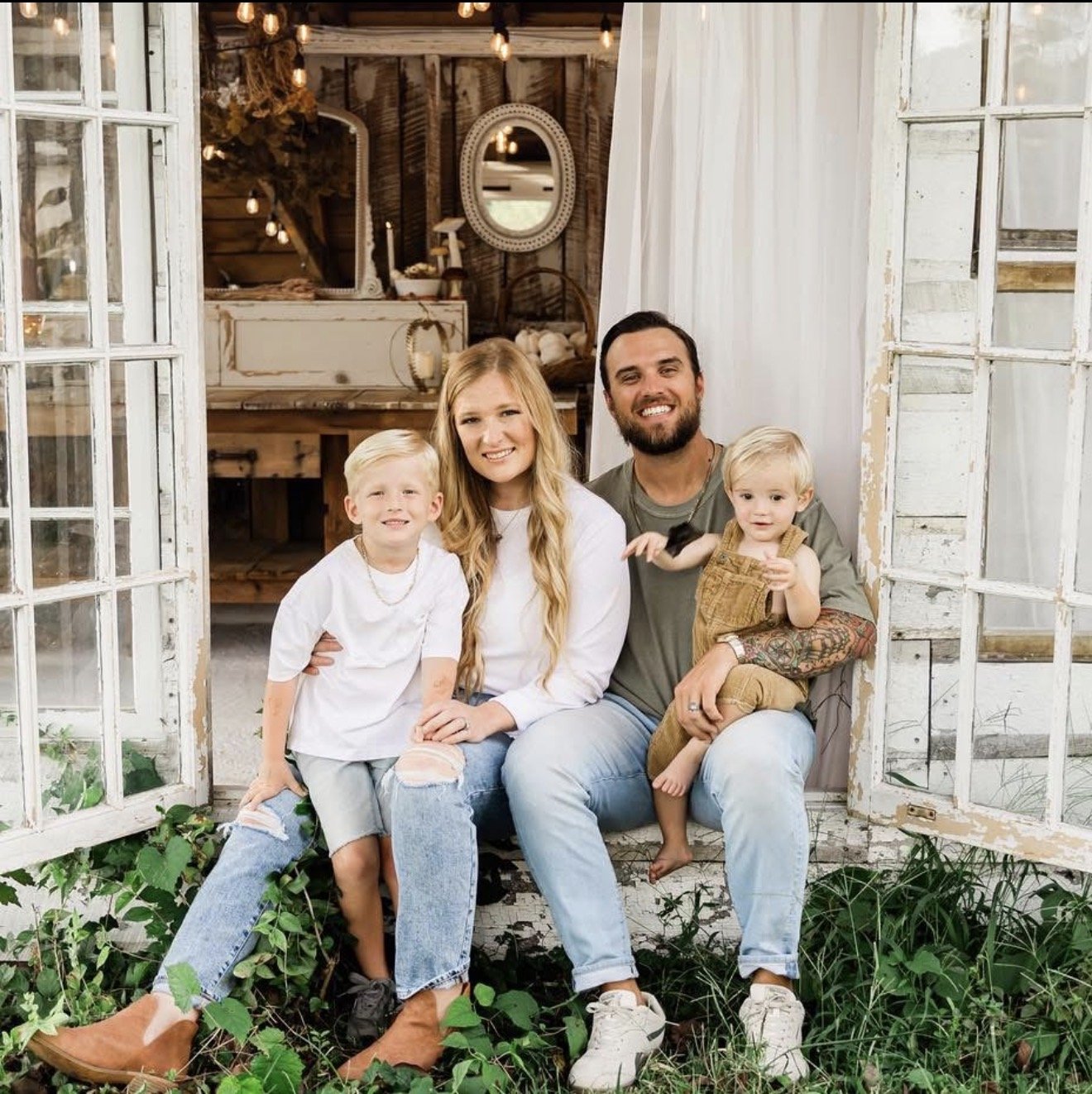 A family of four posing on a rustic porch, with white weathered doors and greenery at their feet, smiling at the camera.