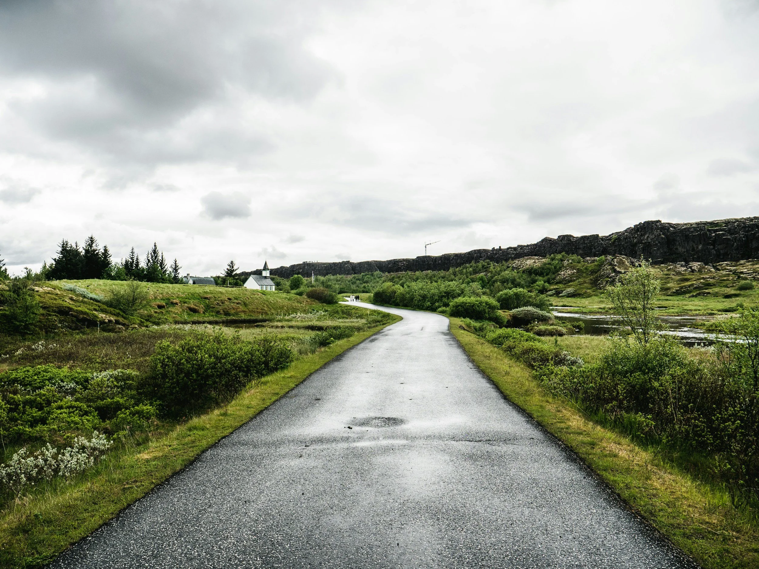 A winding paved road through green fields and trees with a small white church with a bell tower in the distance, under a cloudy sky.