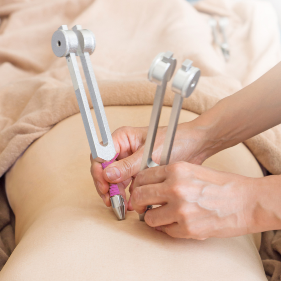 Practitioner holding two weighted tuning forks against a client’s abdomen during a sound therapy session.