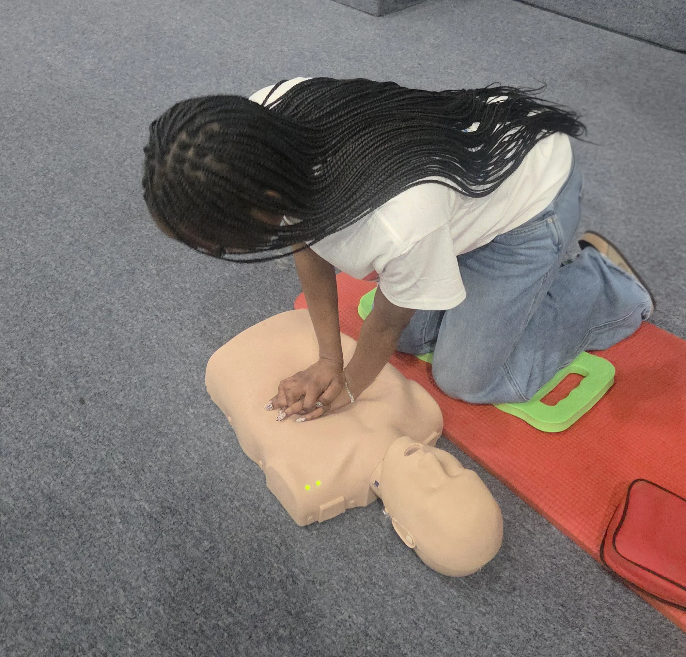 Person lying on red and green mat on a gray carpeted floor, wearing blue jeans and a white shirt, with braided black hair, resting their head on a mannequin head.