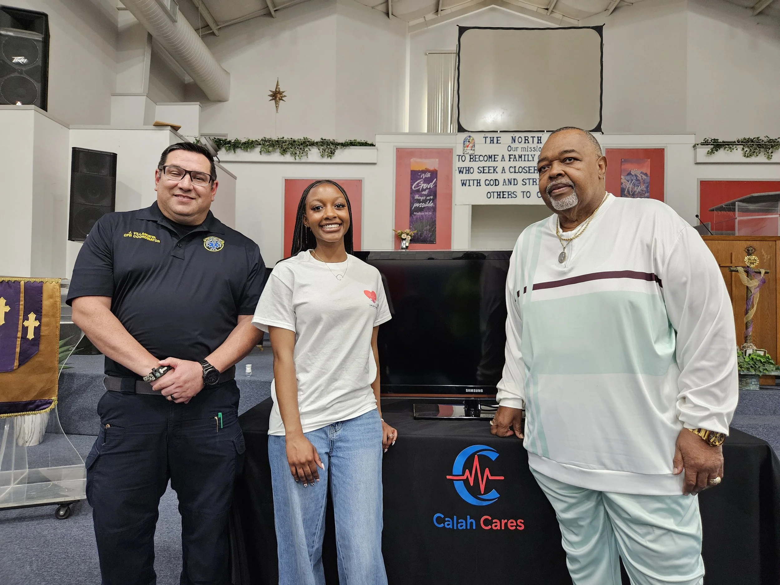 Three people posing inside a church. A woman in the middle and two men on either side. The man on the left wears glasses and a black shirt with a badge. The woman in the middle wears a white T-shirt with a red heart. The man on the right wears a striped shirt with gold chains. The background includes church banners, a speaker, and a sign that reads 'The North' with a mission statement related to faith and community.