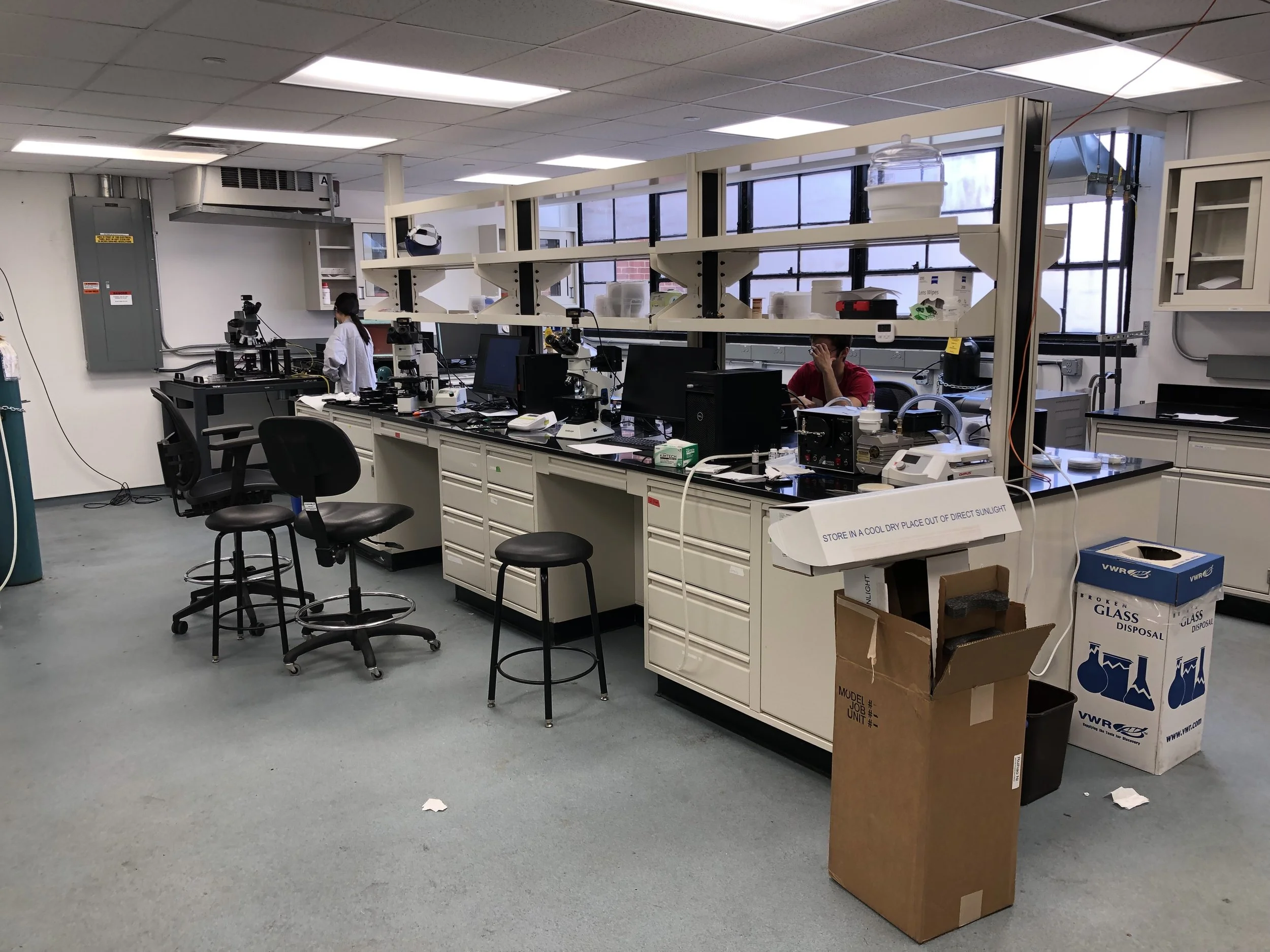 Laboratory room with workbenches, microscopes, computers, and shelves. A woman in a lab coat and a man wearing a red shirt are working amidst various scientific equipment and supplies.