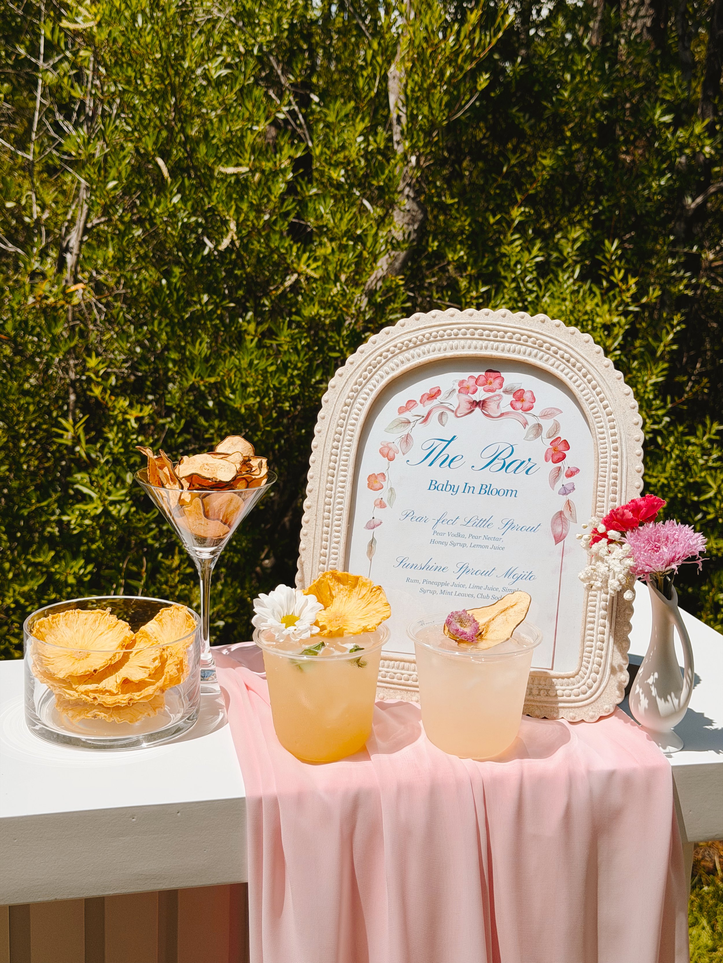 A display of beverages curated by The Salty Rim, with two fruity drinks, pineapple garnishes, and a menu board labeled 'The Bar' at an outdoor baby shower event with a pink tablecloth and floral decorations.