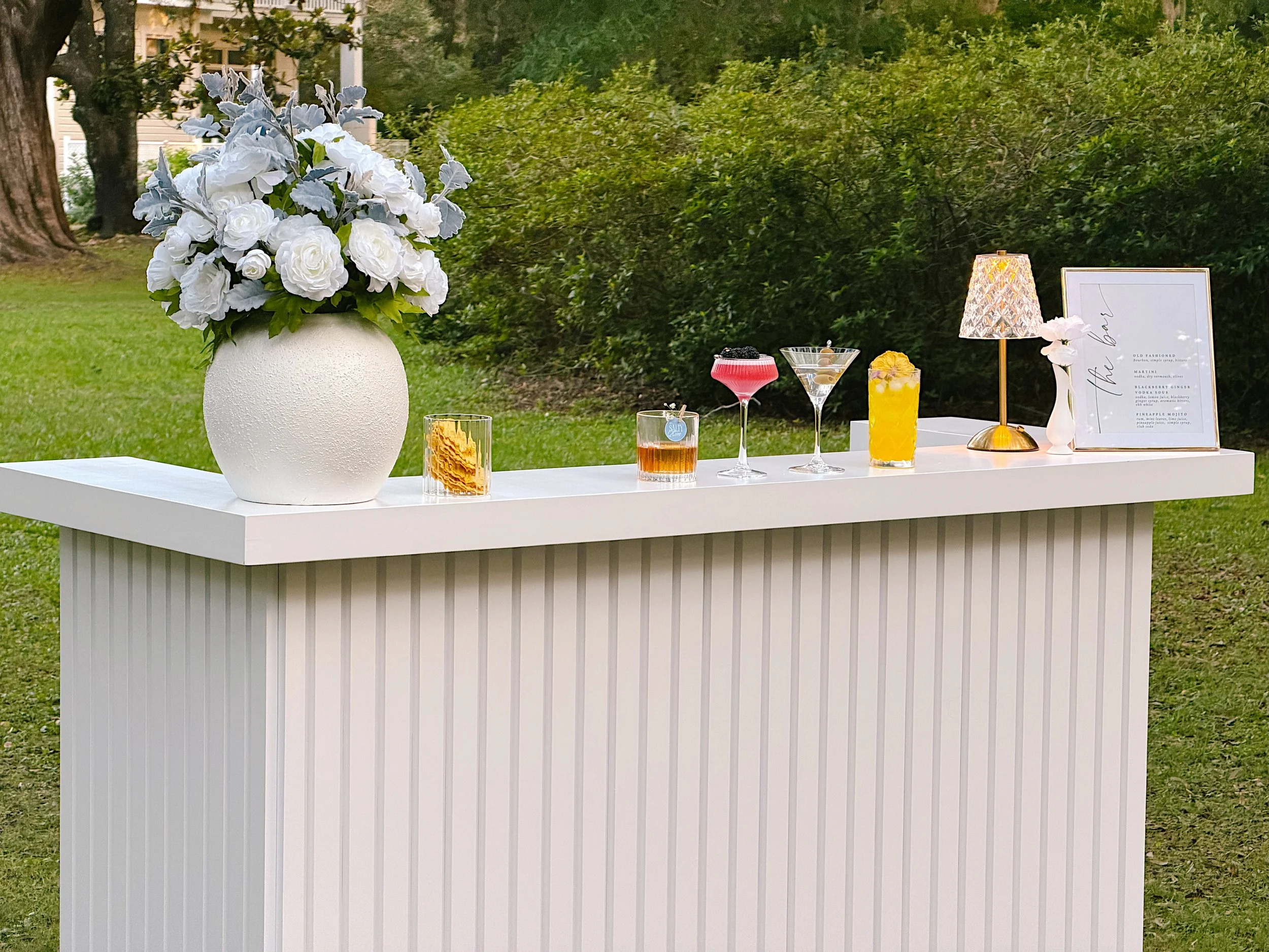A white portable bar from The Salty Rim with a large floral arrangement in a textured white vase, colorful cocktails, a lamp, and framed artwork, set outdoors with trees and bushes in the background.