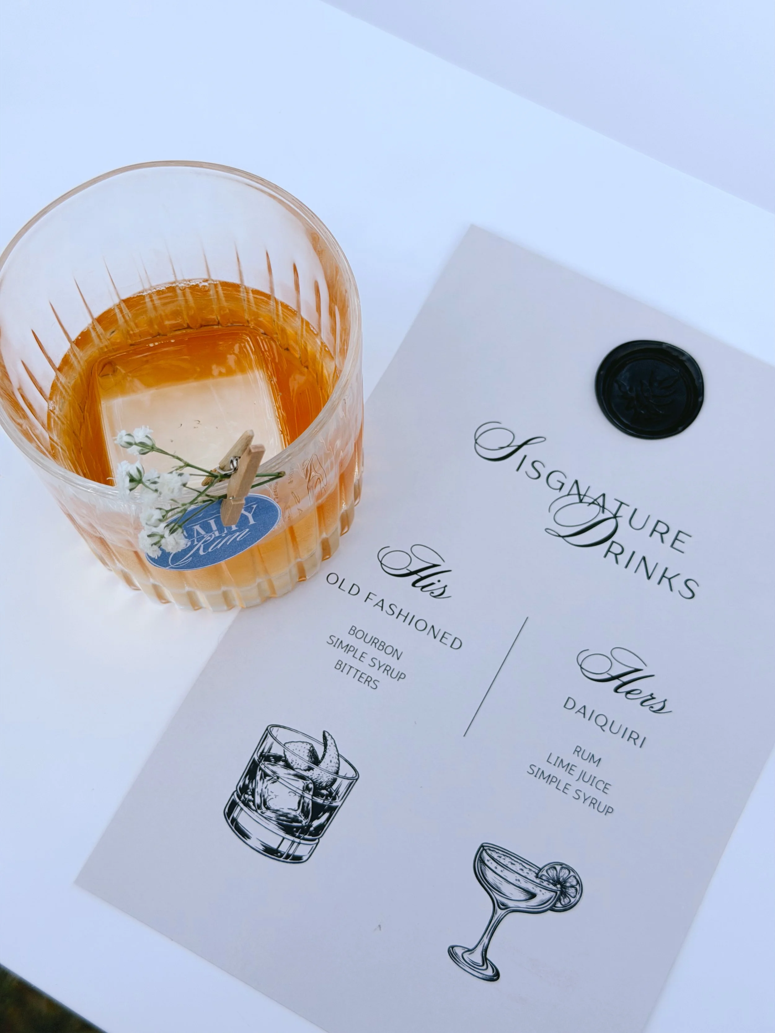 A glass with an old fashioned whiskey cocktail with a small white flower decoration and a clothespin holding The Salty Rim logo, placed on a white paper menu that lists various cocktails.