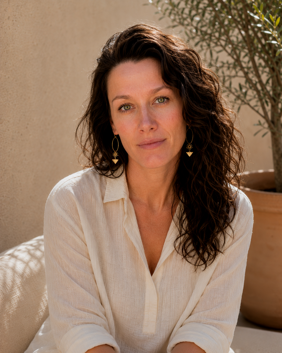 A woman with wavy brown hair and blue eyes looking at the camera, wearing a cream-colored shirt and star and heart-shaped earrings, sitting outdoors near a large potted plant.