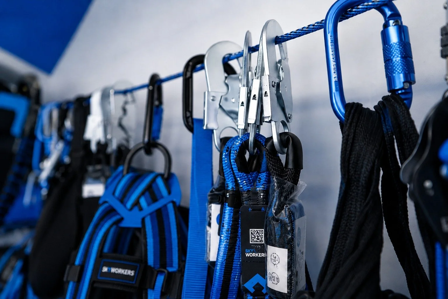 Verschiedene Klettergurte und Karabiner hängen an einem blauen Stahlseil an einer Wand.