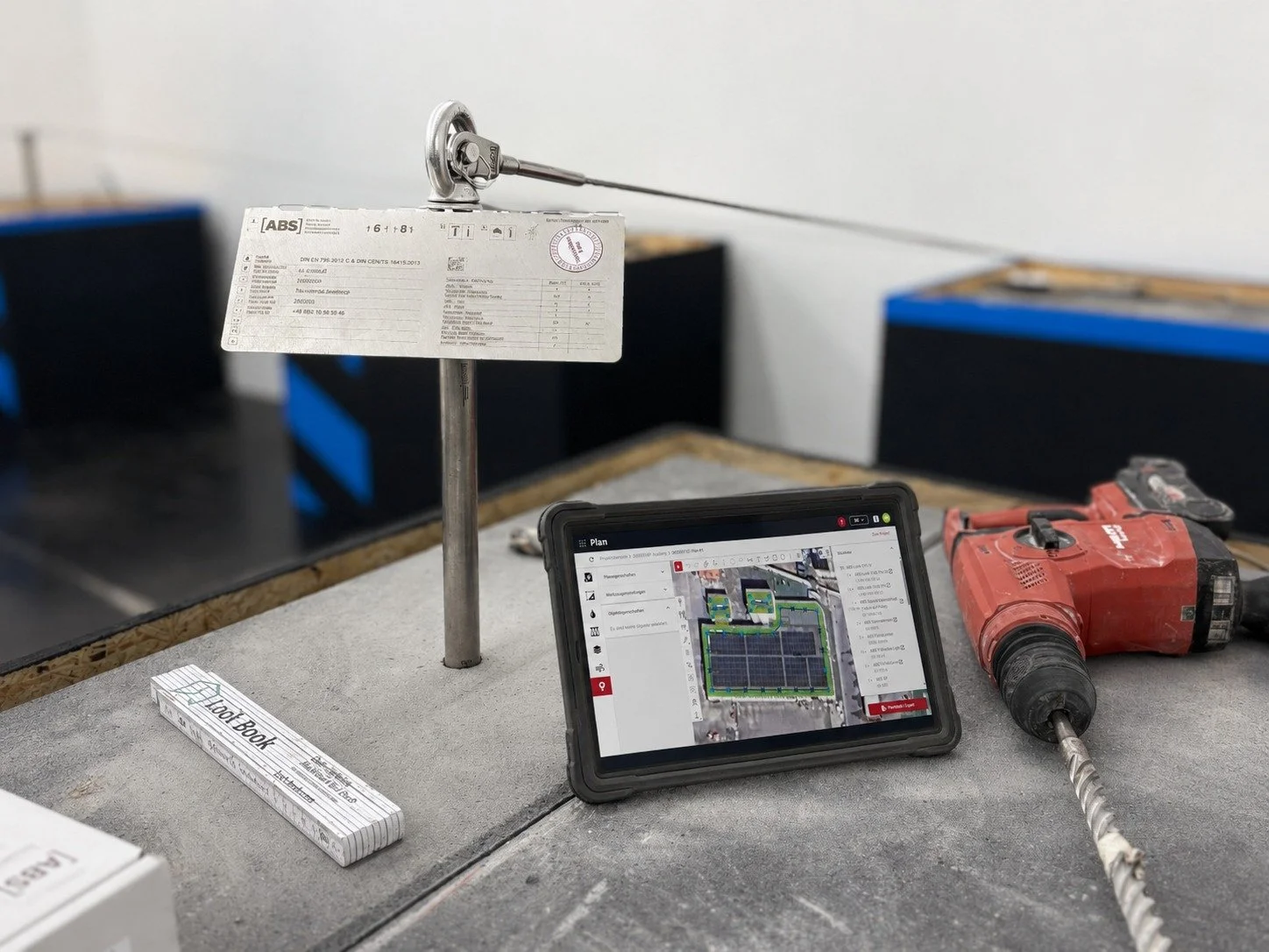 Auf einer Baustelle: Ein Tablets mit Bauplan, ein rotes Bohrgerät, ein Messgerät, ein A4-Block und eine Werkzeugeinheit mit Karton. Im Hintergrund sind schwarze und blaue Gegenstände.