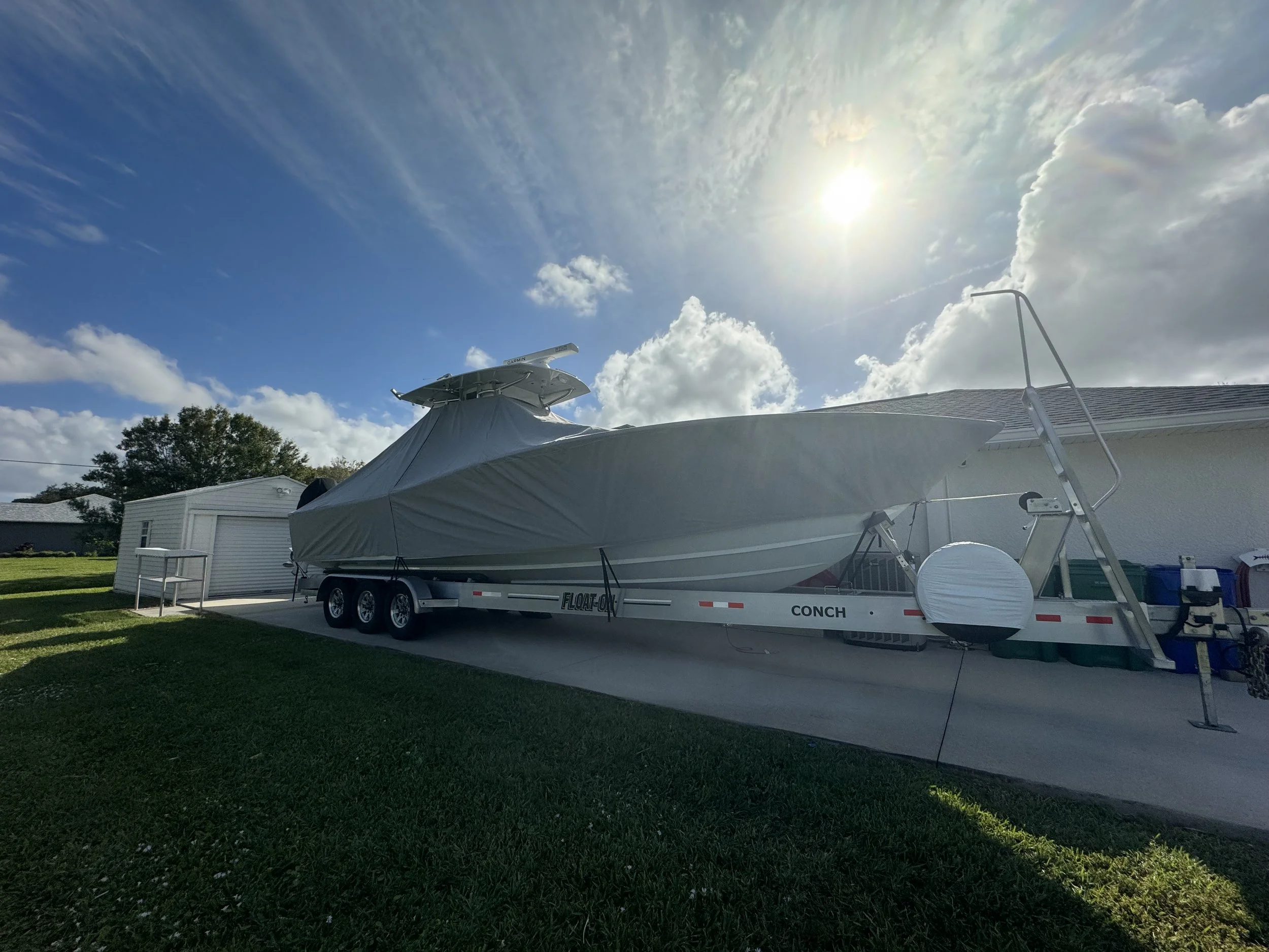 A boat on a trailer parked in a backyard with a white shed nearby. The sky is partly cloudy with the sun shining through, creating a bright atmosphere.