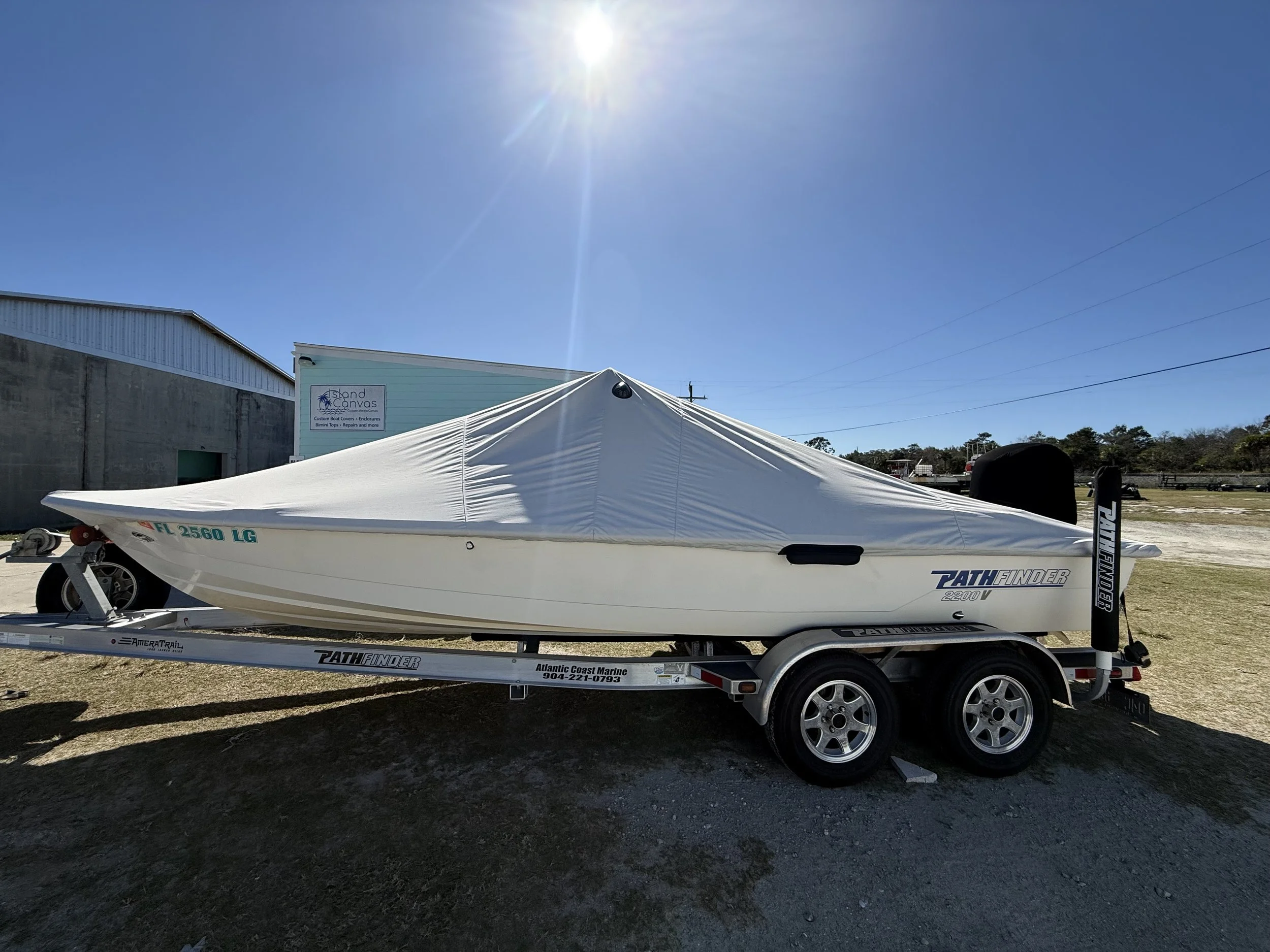 A white boat covered with a white tarp on a trailer, parked outdoors under a clear blue sky with bright sunlight.
