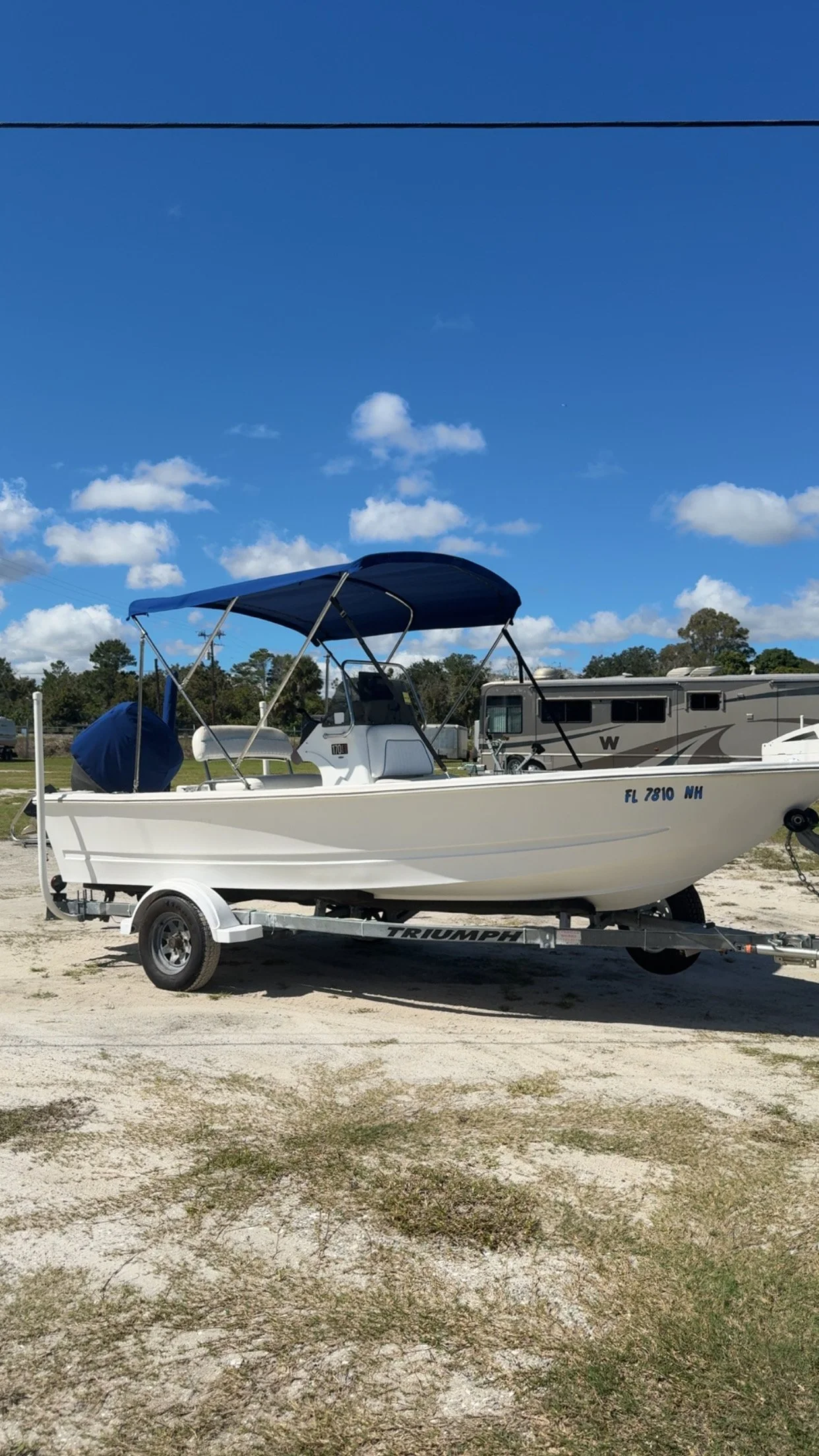 A white boat on a trailer in a sandy outdoor area with grass, under a blue sky with scattered clouds and power lines. Behind the boat are trees and RVs.