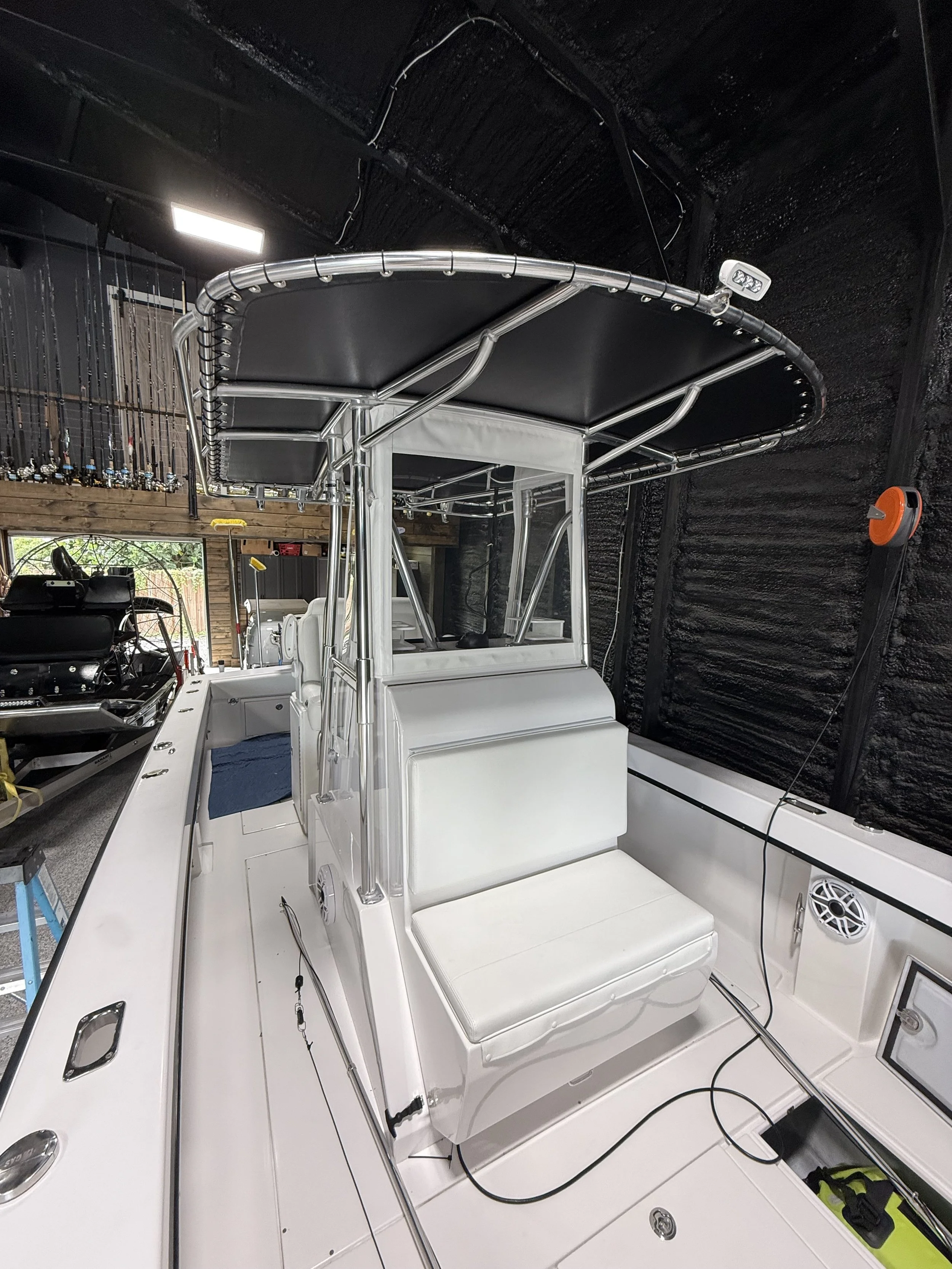 A white fishing boat with a T-top shade structure inside a workshop or garage. The boat has binoculars mounted on the T-top and fishing rods on the side, with electronics and equipment stored on the deck.