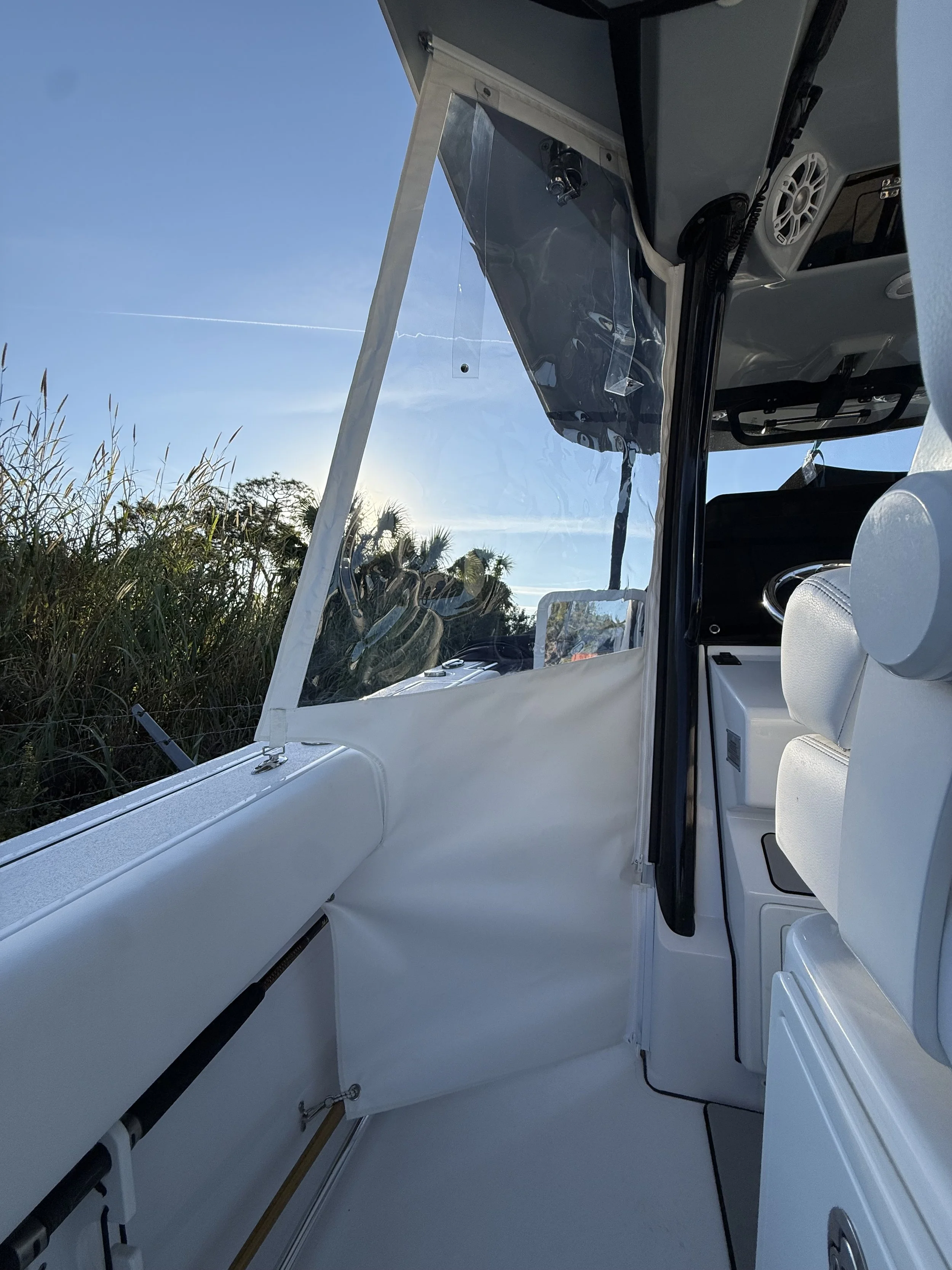 Interior of a small white boat with seating, controls, and a clear windshield, set outdoors with a grassy area and trees visible through the windshield and to the side, with a clear sky overhead.