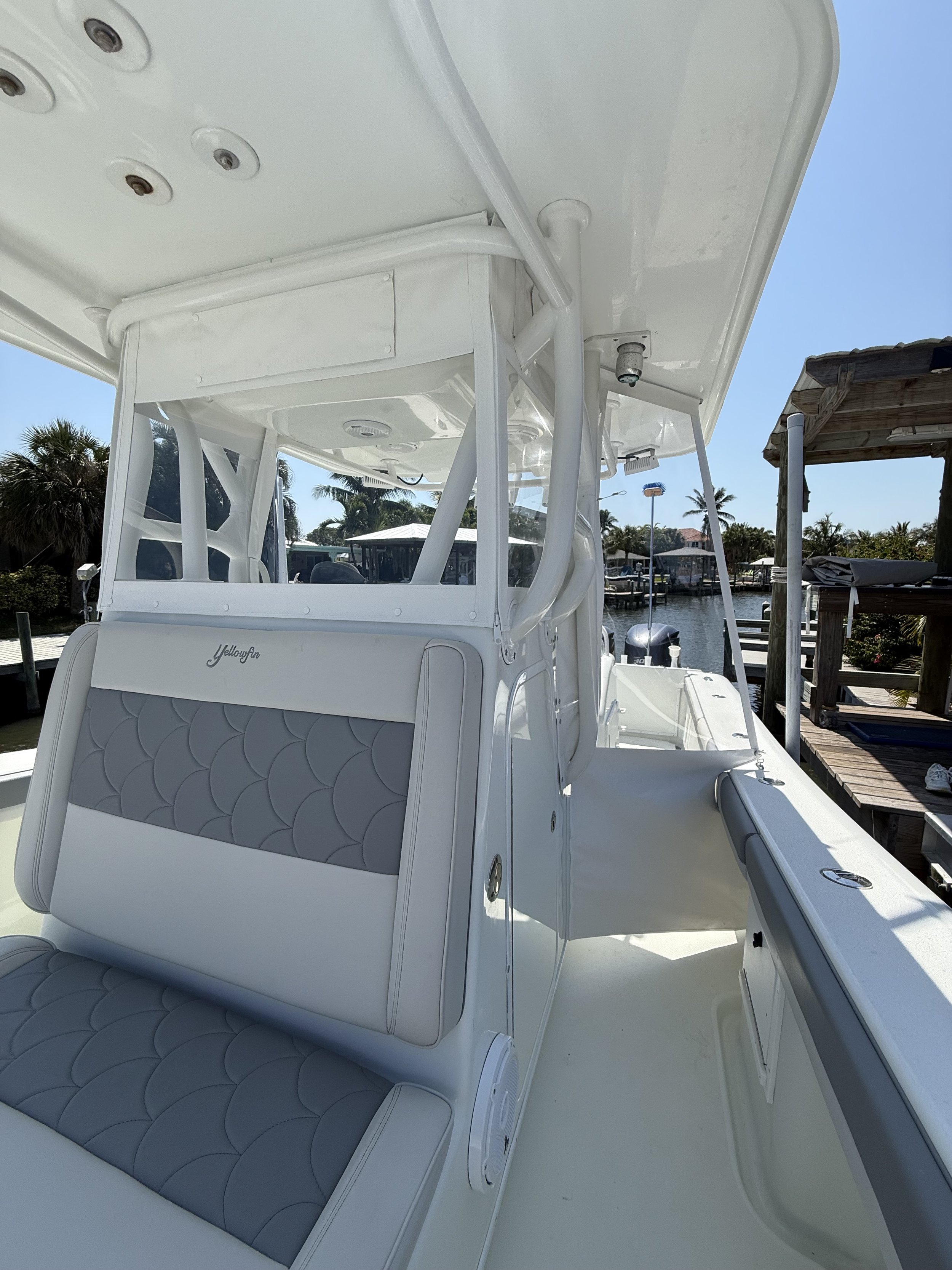 View of a white yacht boat with gray cushioned seating, a canopy, and boat controls, docked at a marina with palm trees, water, and other boats in the background.