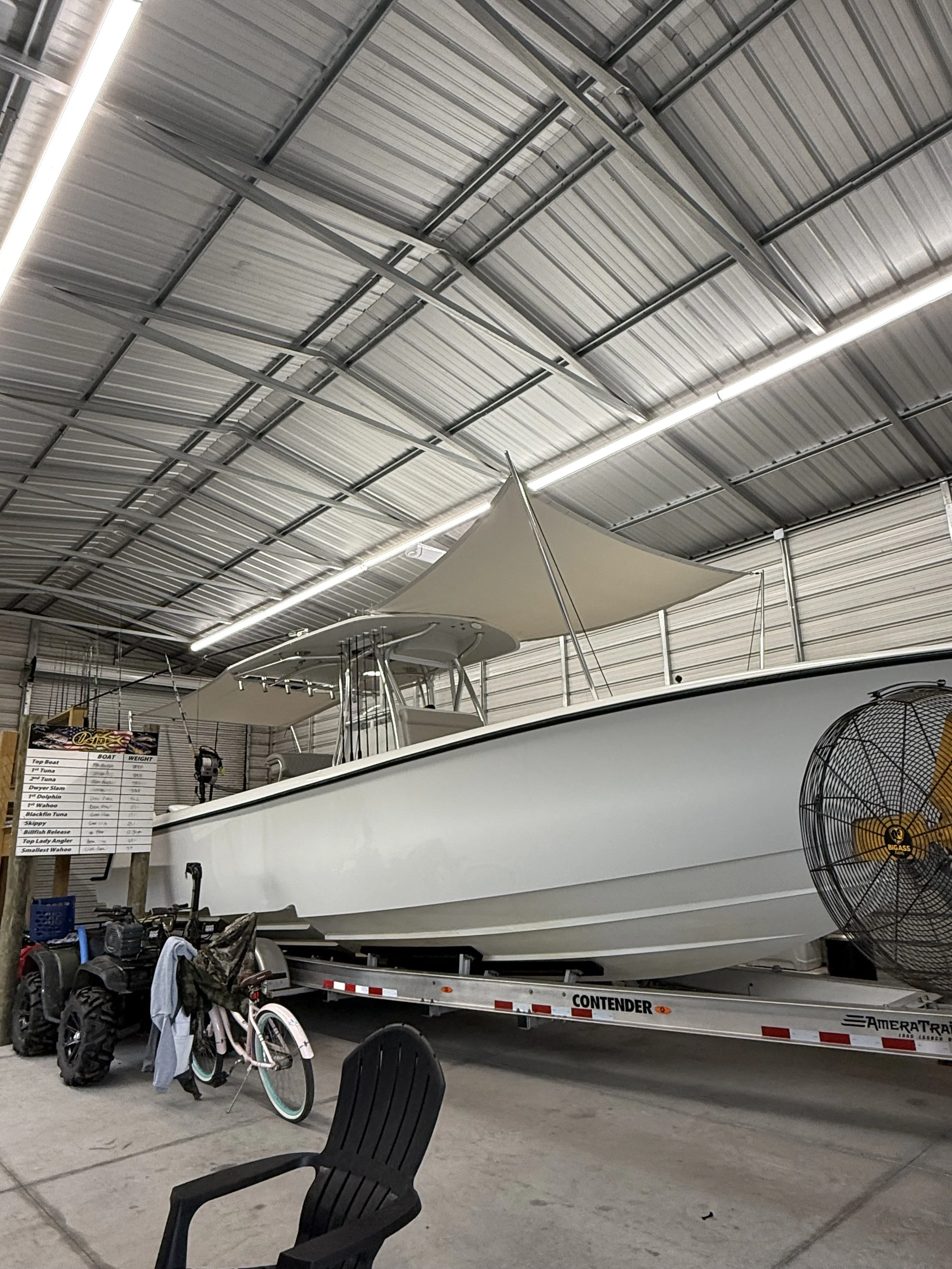 An indoor storage facility with a boat on a trailer, a bicycle, a large fan, a black chair, and a small table.
