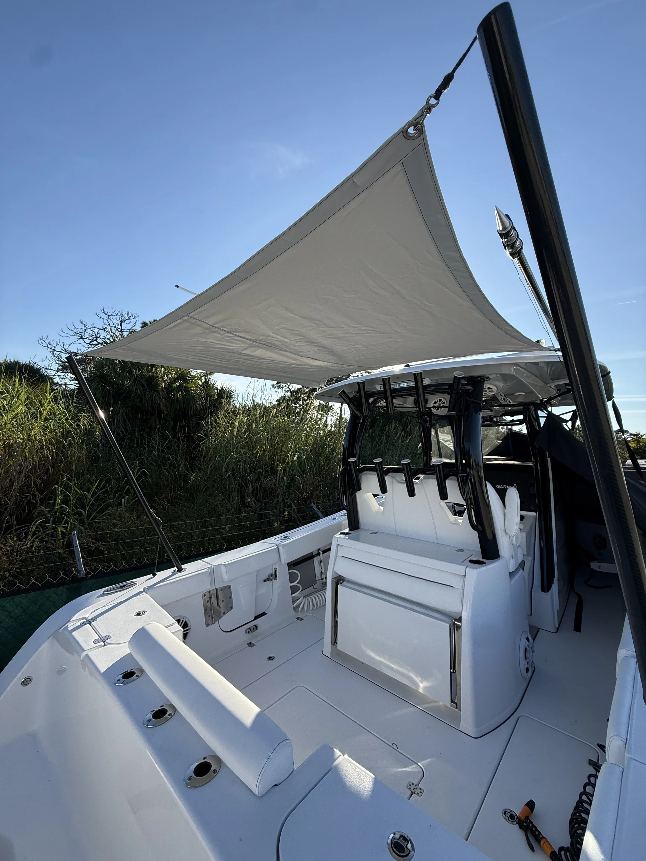 A boat with a shade canopy, fishing rods, and a seating area, situated outdoors near green vegetation and a blue sky.
