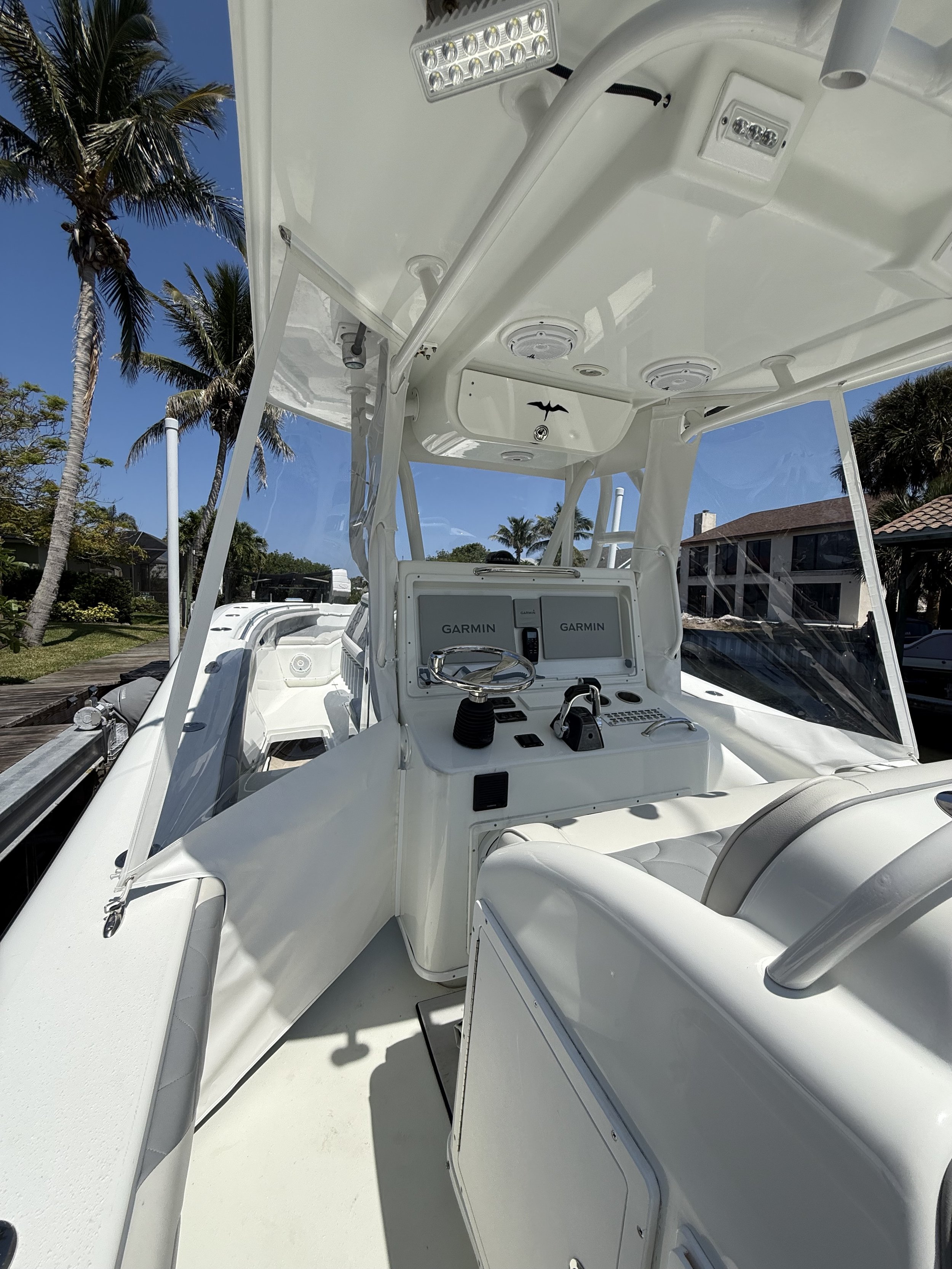 The image shows the interior of a white boat with two Garmin electronic screens, a steering wheel, and various controls. The boat's ceiling has built-in lights and speakers. Outside the boat, there are trees, houses, and a clear blue sky.