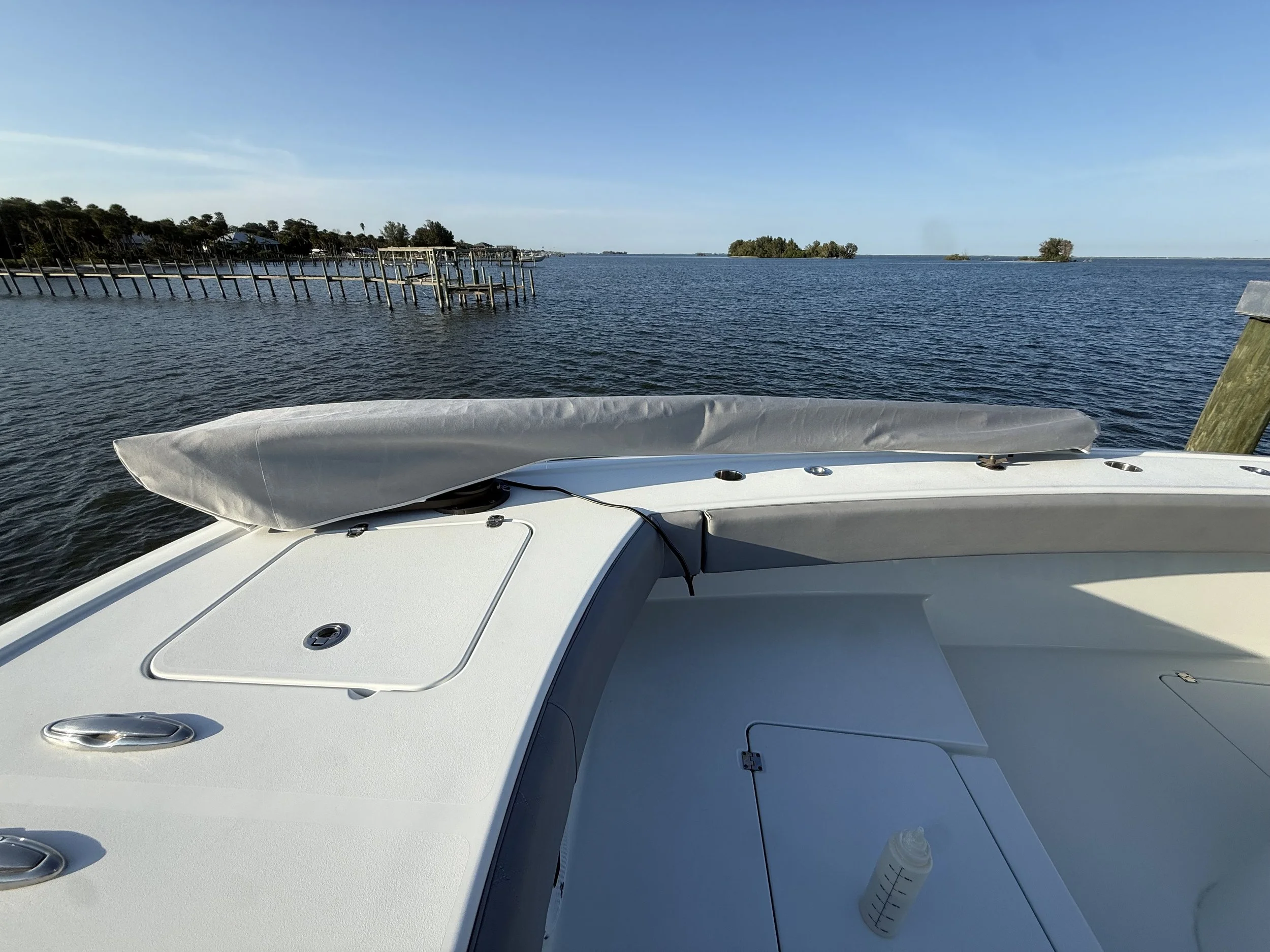 View from the deck of a white boat on a river or lake, with calm water, a dock with multiple boat slips on the left, small islands in the distance, and a clear blue sky.