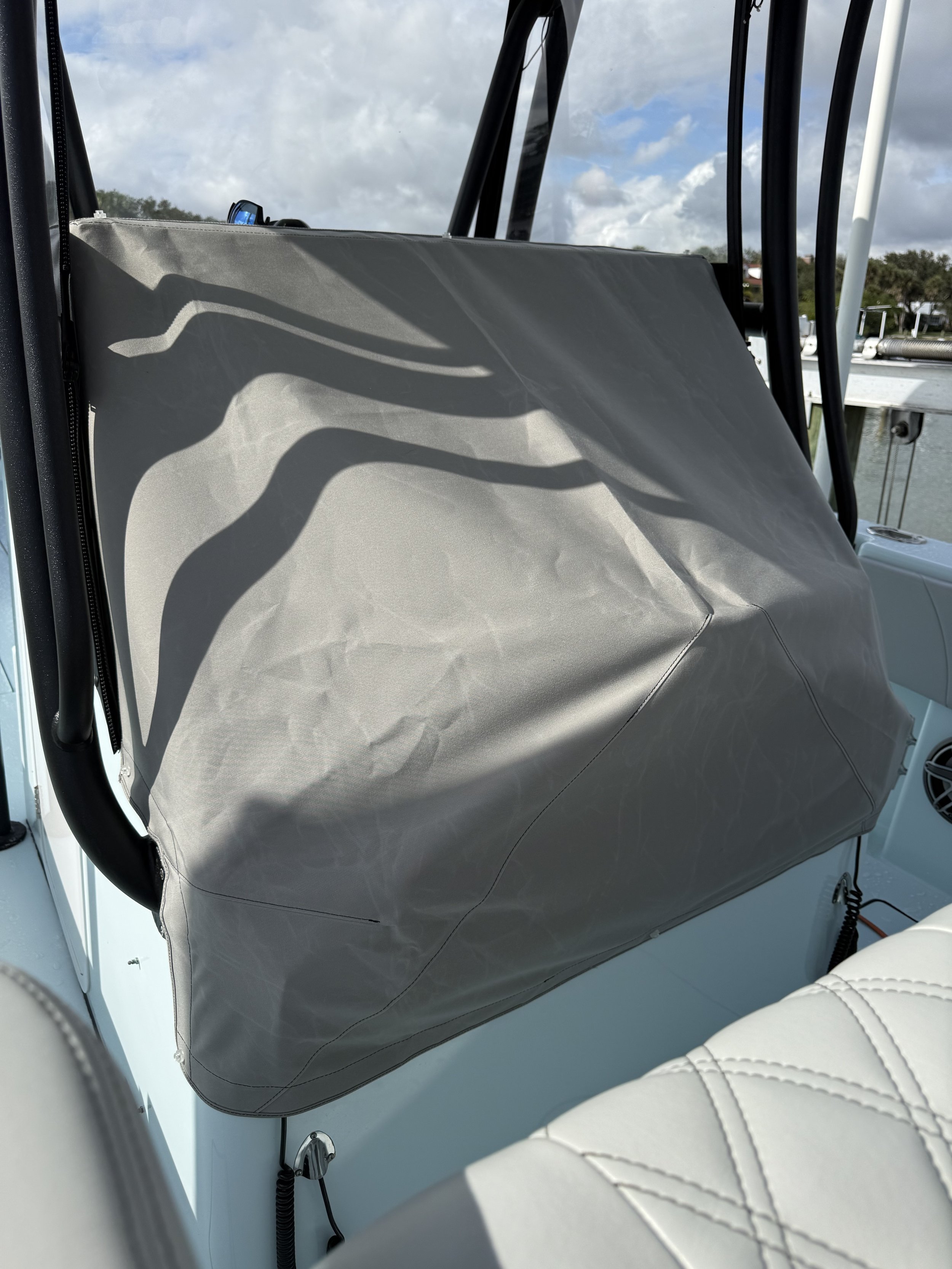 The image shows a boat's interior with a gray cover on the back seat and a white cushioned seat in the foreground. There are black metal bars and a boat's control panel visible, with water and cloudy sky in the background.