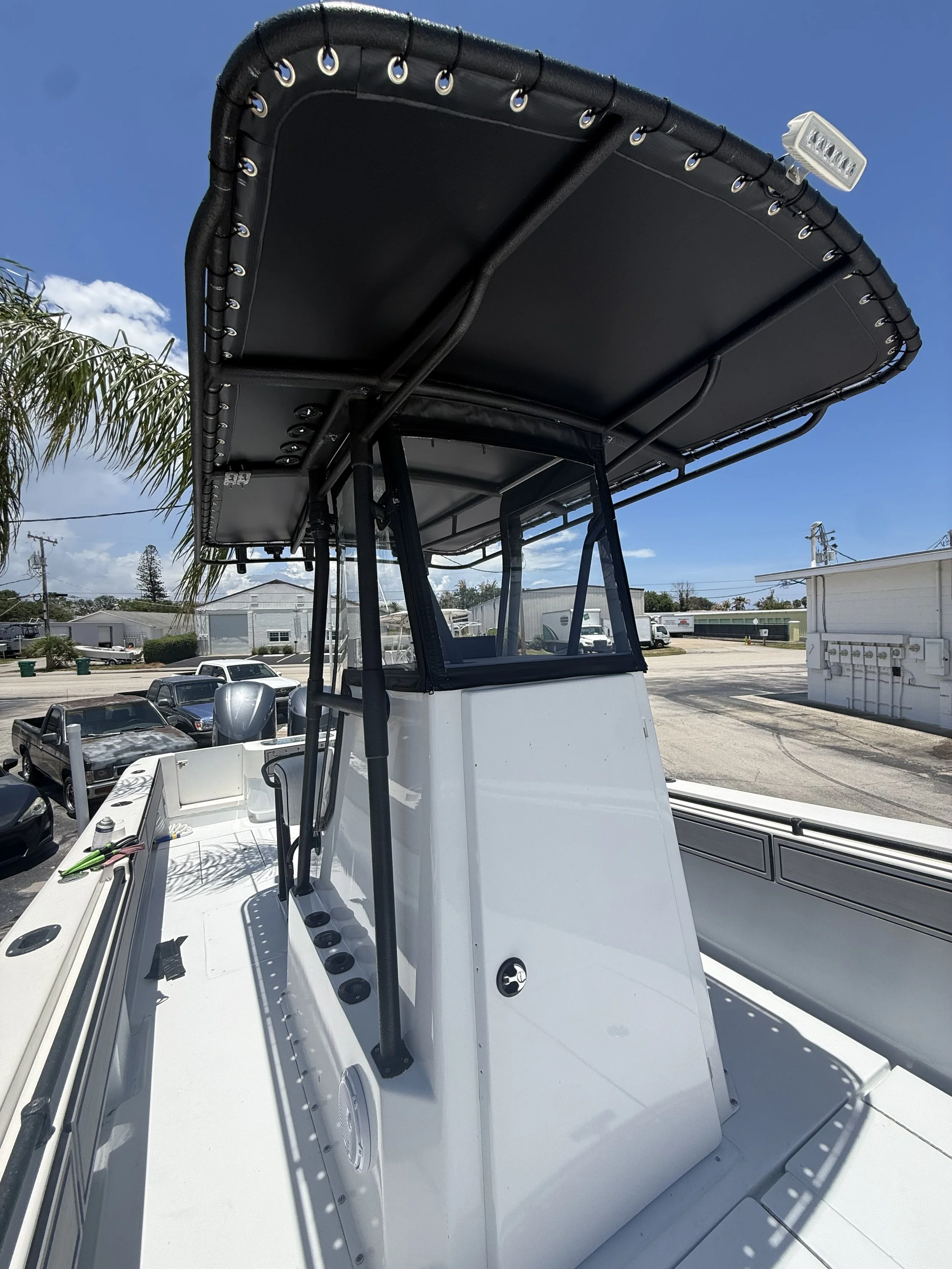 A white boat with a black canopy top, held up by metal supports, is parked outdoors. In the background, there are several parked vehicles, a building, power lines, and palm trees under a blue sky with few clouds.