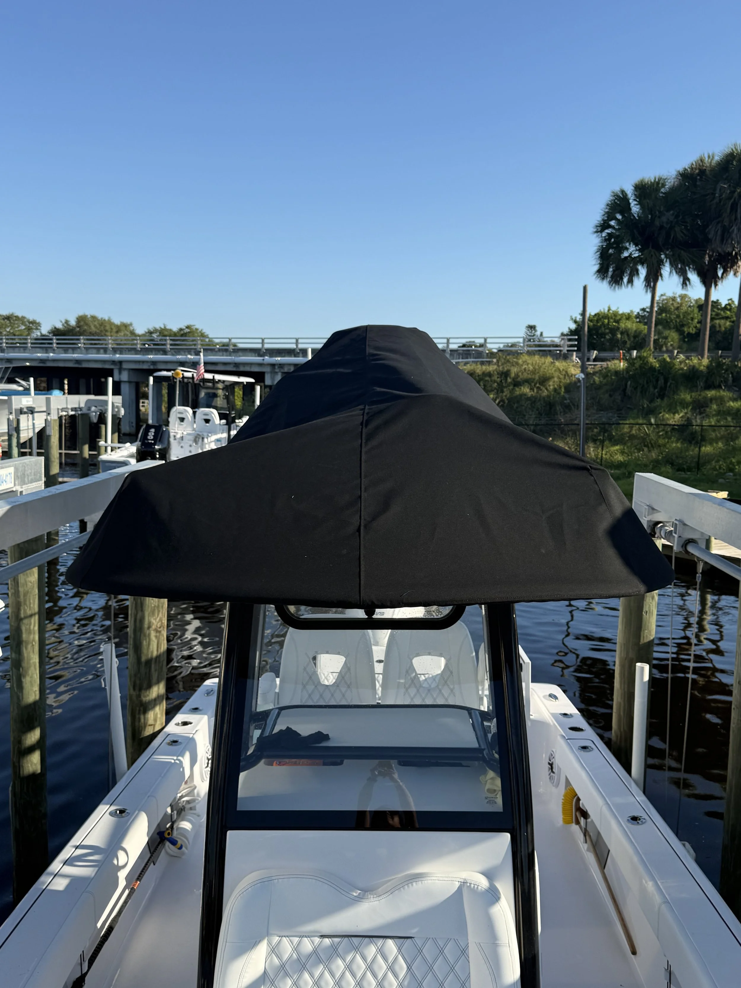 View of a boat docked at a marina with a large black cover over the helm, white seating, water, and palm trees in the background.