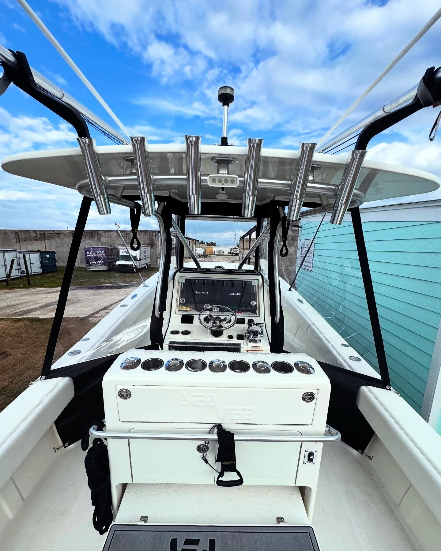 The interior of a white boat showing the helm with a GPS display, steering wheel, and control buttons, with fishing rods mounted above and a view of a blue sky with clouds overhead.
