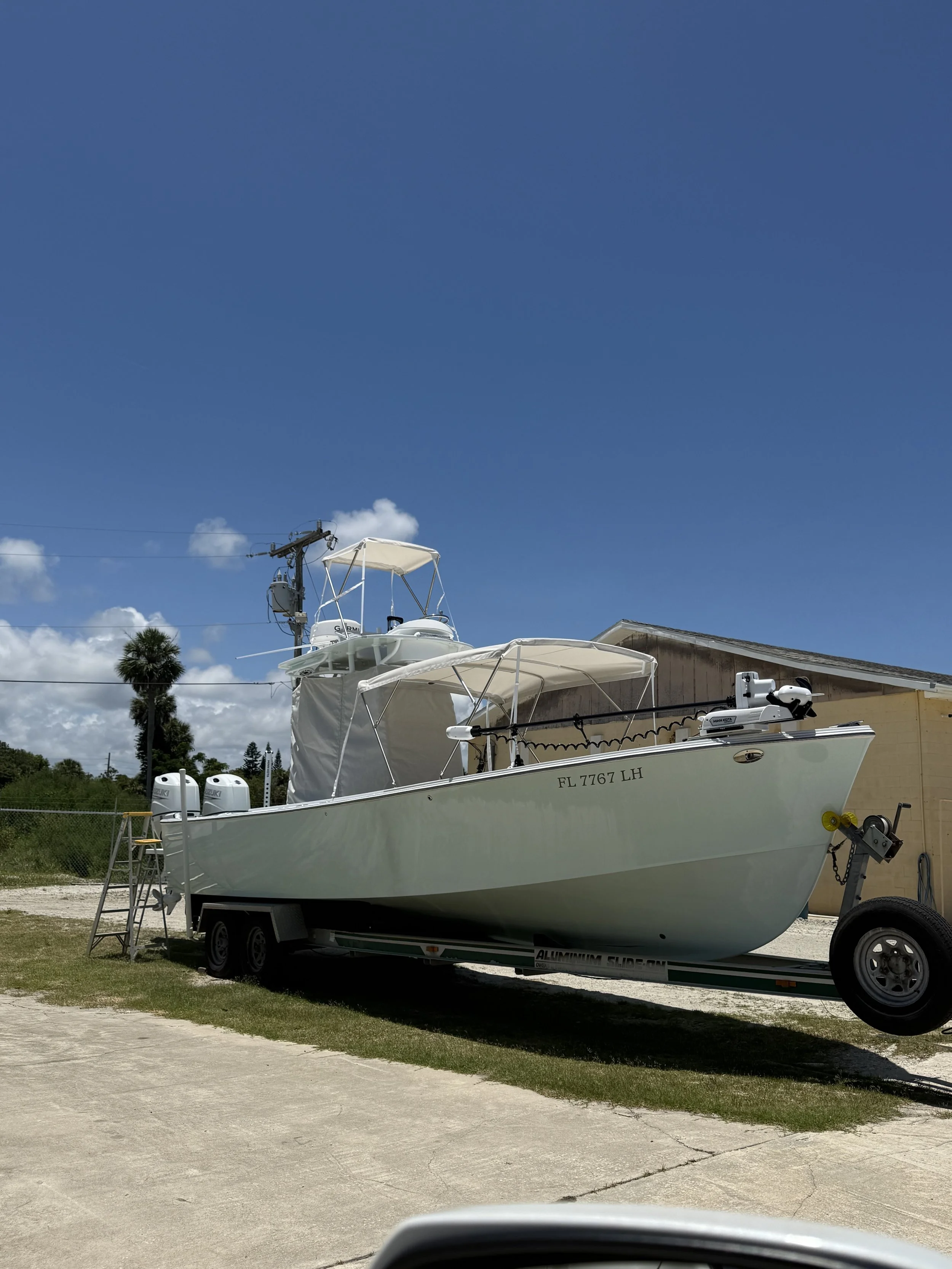 A white boat on a trailer parked on a driveway next to a building with a yellow wall. The boat has a canopy and is equipped with outboard motors. There are sandals on a step ladder next to the boat. A utility pole with power lines and some trees are 