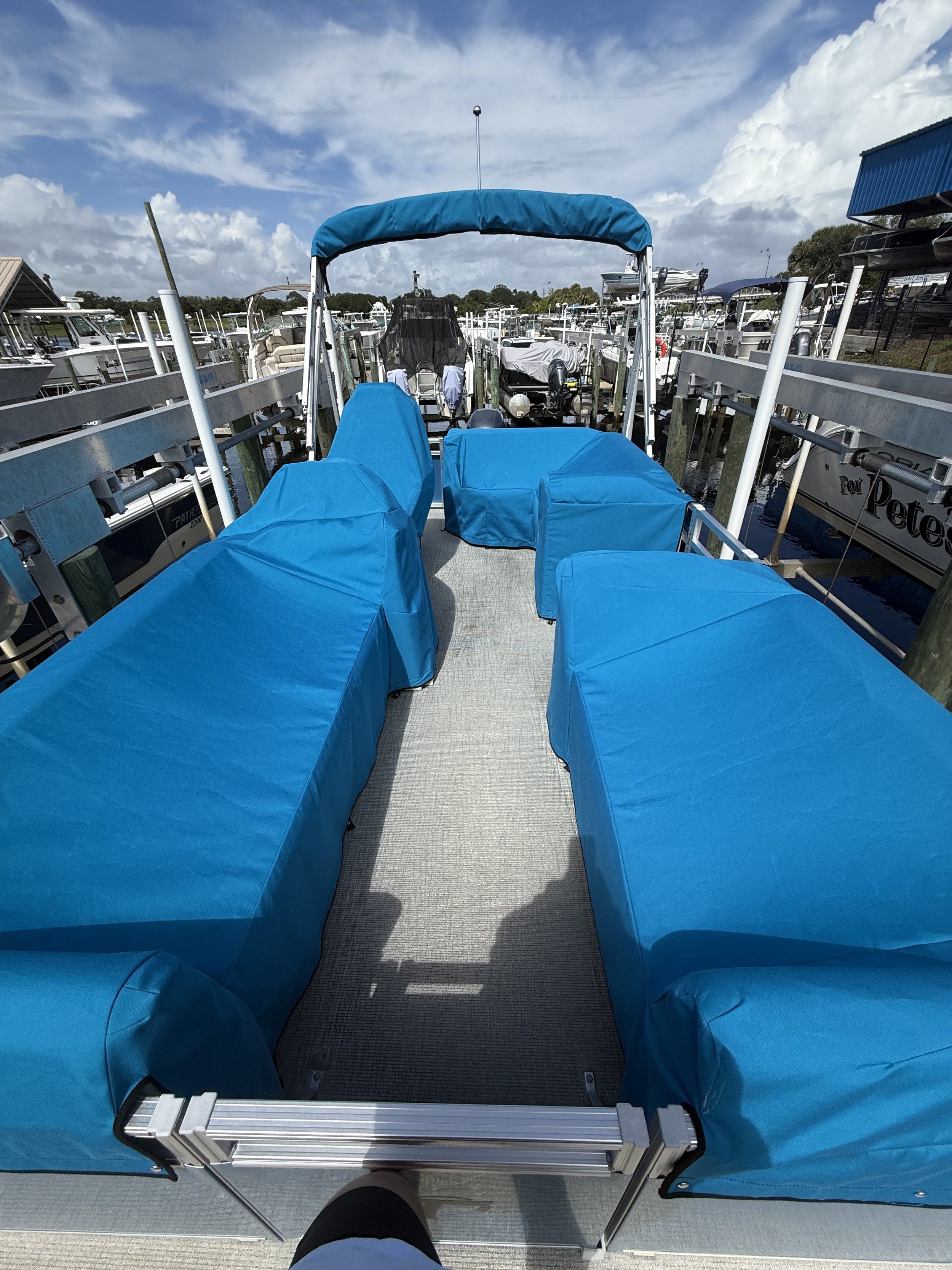 View of a boat deck with four blue covered seats, a black foot, and a walkway leading toward the back of the boat. In the background, there are multiple boats docked at a marina under a partly cloudy sky.