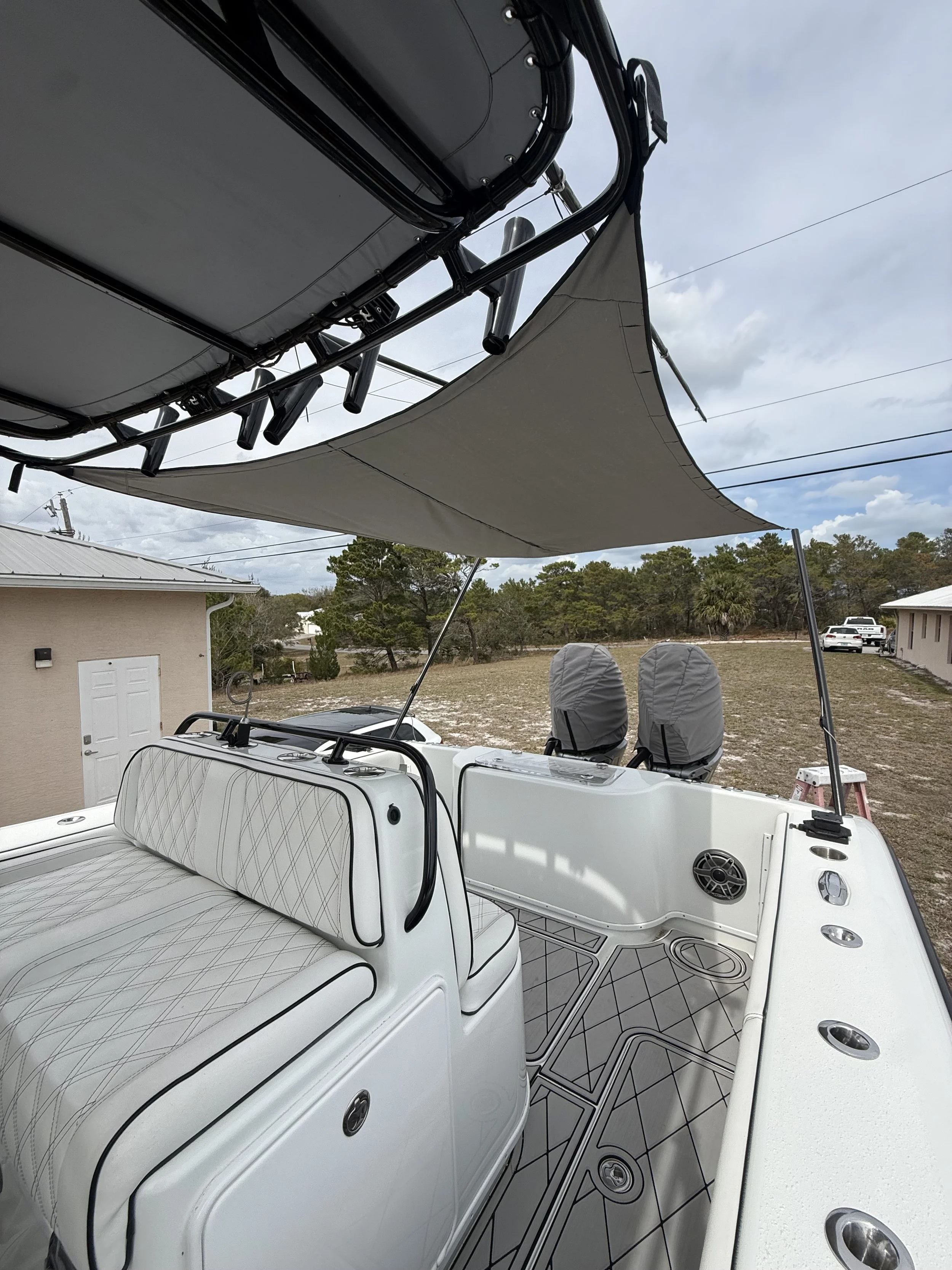 View of the interior and roof of a white boat with cushioned seating, featuring a shaded canopy and two outboard motors, with a background of a house, parked cars, and trees under a cloudy sky.