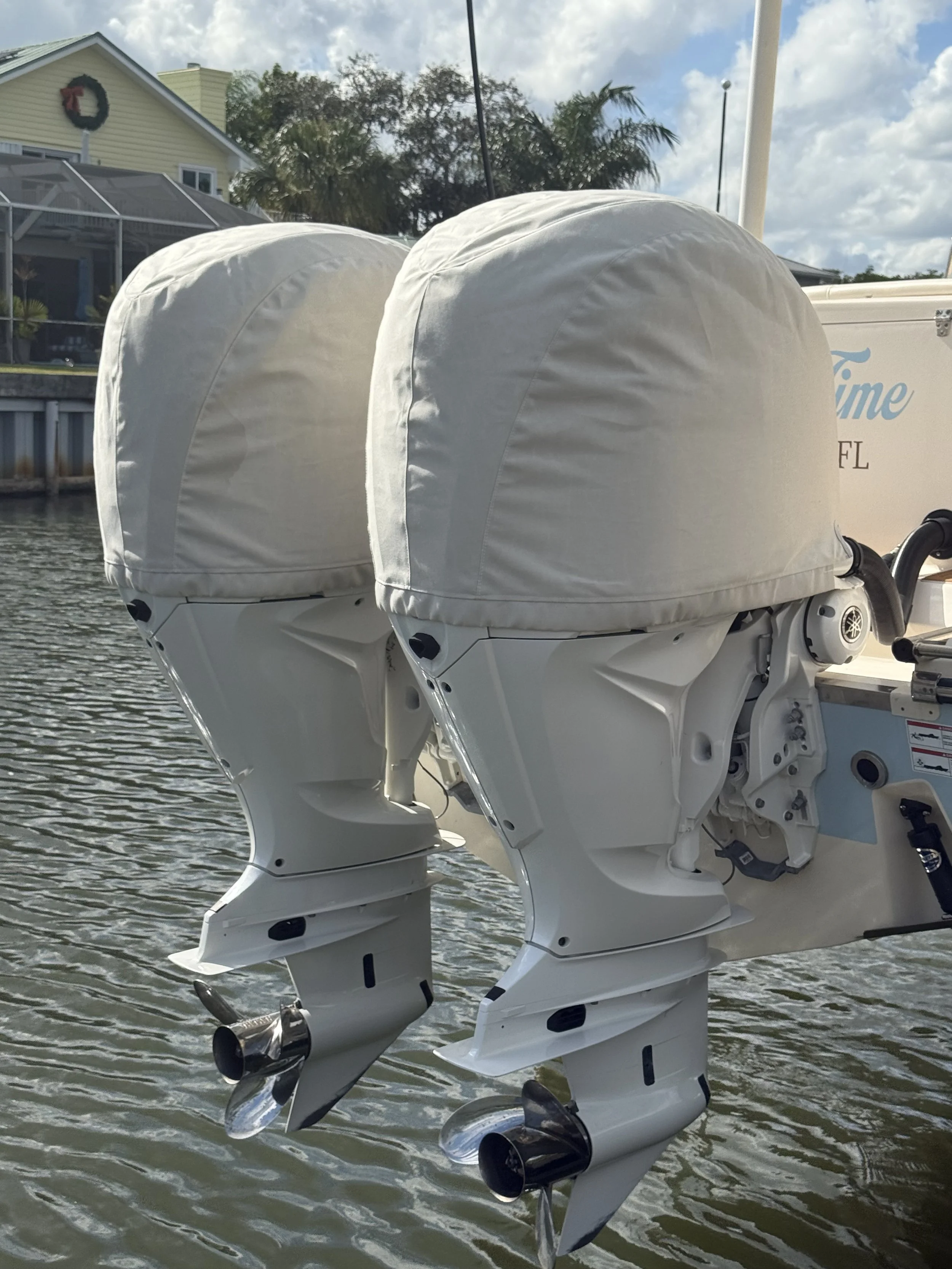 Two white covered boat engines mounted on the back of a boat on the water with houses and trees in the background.