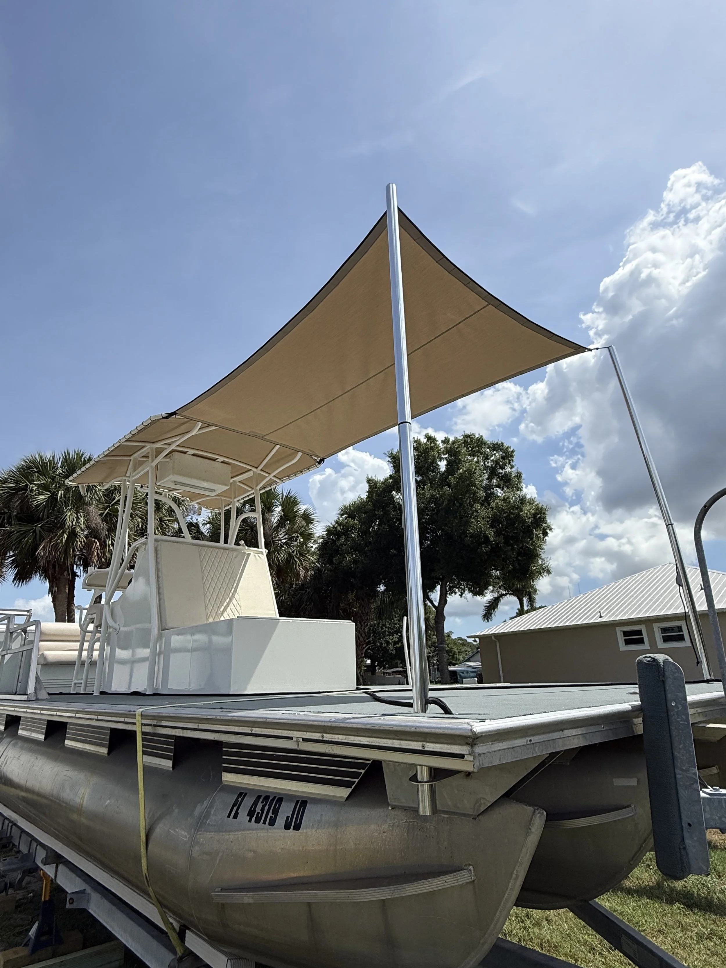 A pontoon boat with a beige canopy on a trailer in an outdoor setting with trees and a building in the background, under a partly cloudy sky.