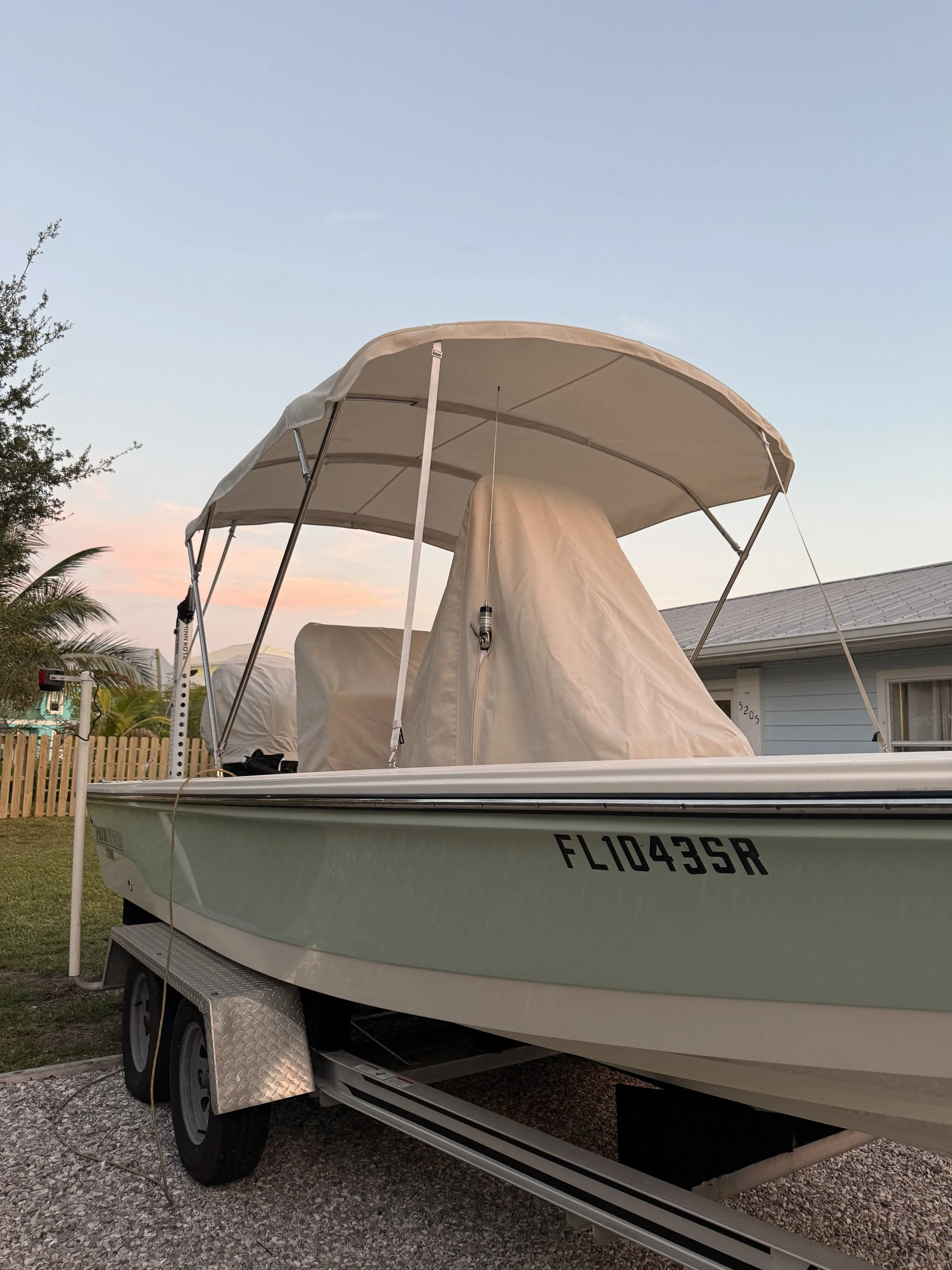 A boat on a trailer in a backyard with a house, a fence, and some trees in the background, under a clear sky during sunset.