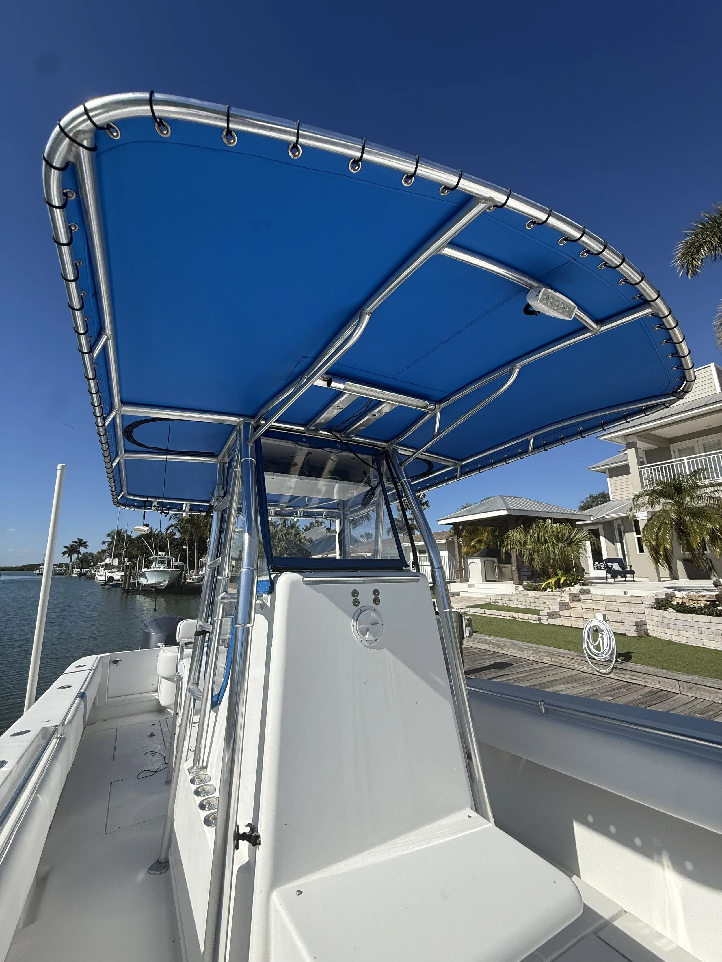 A white boat with a blue canopy docked by a waterfront house with a lawn and palm trees, under a clear blue sky.