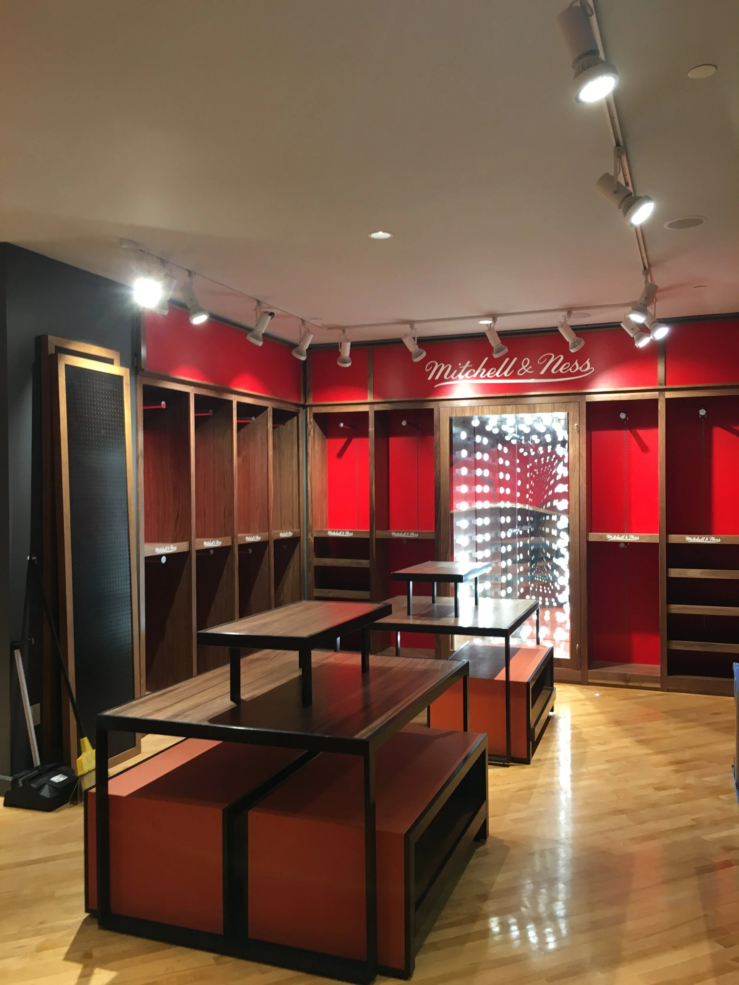 Empty retail space with wooden and black display cases, red and wood wall panels, and a sign that reads 'Mitchell & Ness' in white cursive at the back. Spotlights are on the ceiling, and the floor is hardwood.
