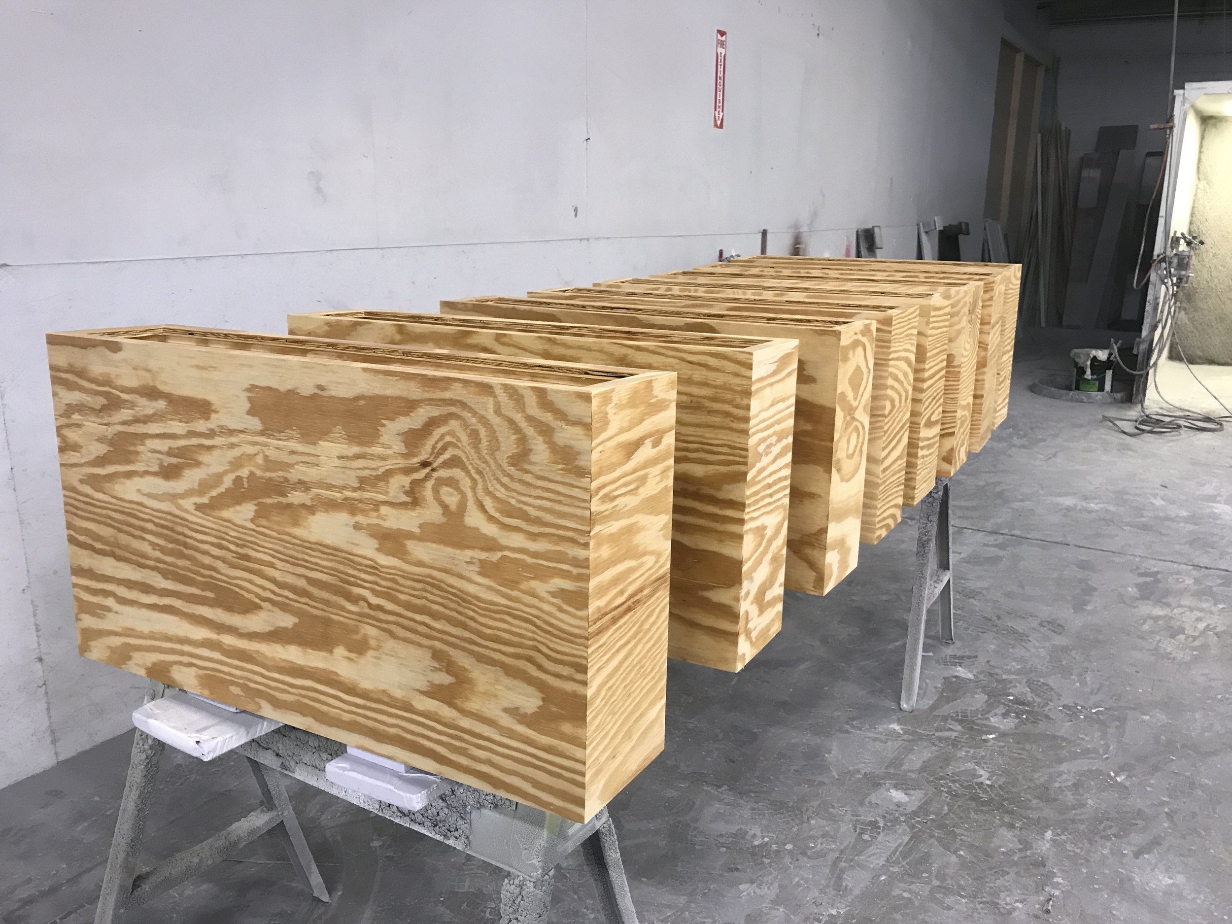 Stacked unfinished wooden boxes in a workshop with a gray concrete floor, white walls, and some tools in the background.