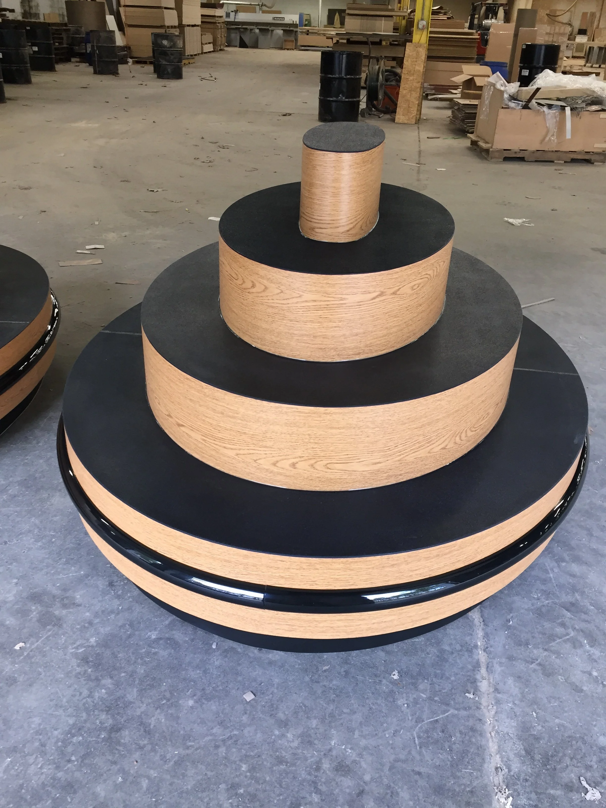 Large circular display stand with three levels made of wood and black surfaces, located in a workshop or warehouse.