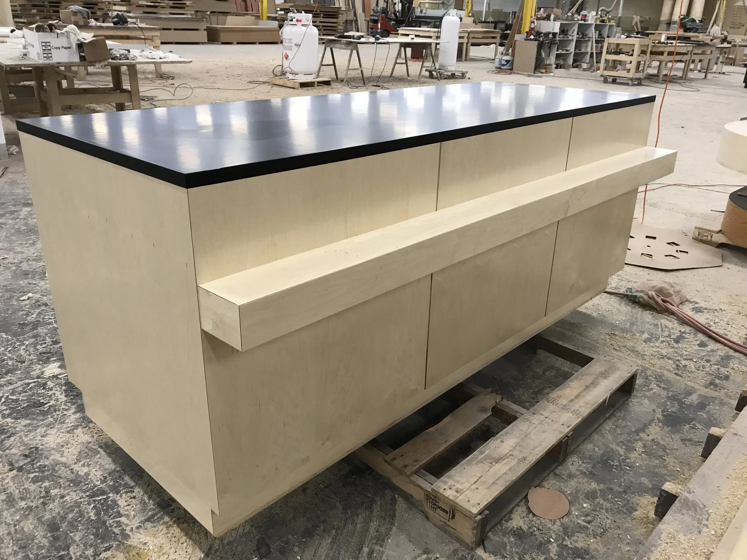 A workspace with a piece of furniture under construction. The furniture has a black top surface and light-colored wooden base. The background shows clutter typical of a woodworking shop, including workbenches, power tools, and supplies.