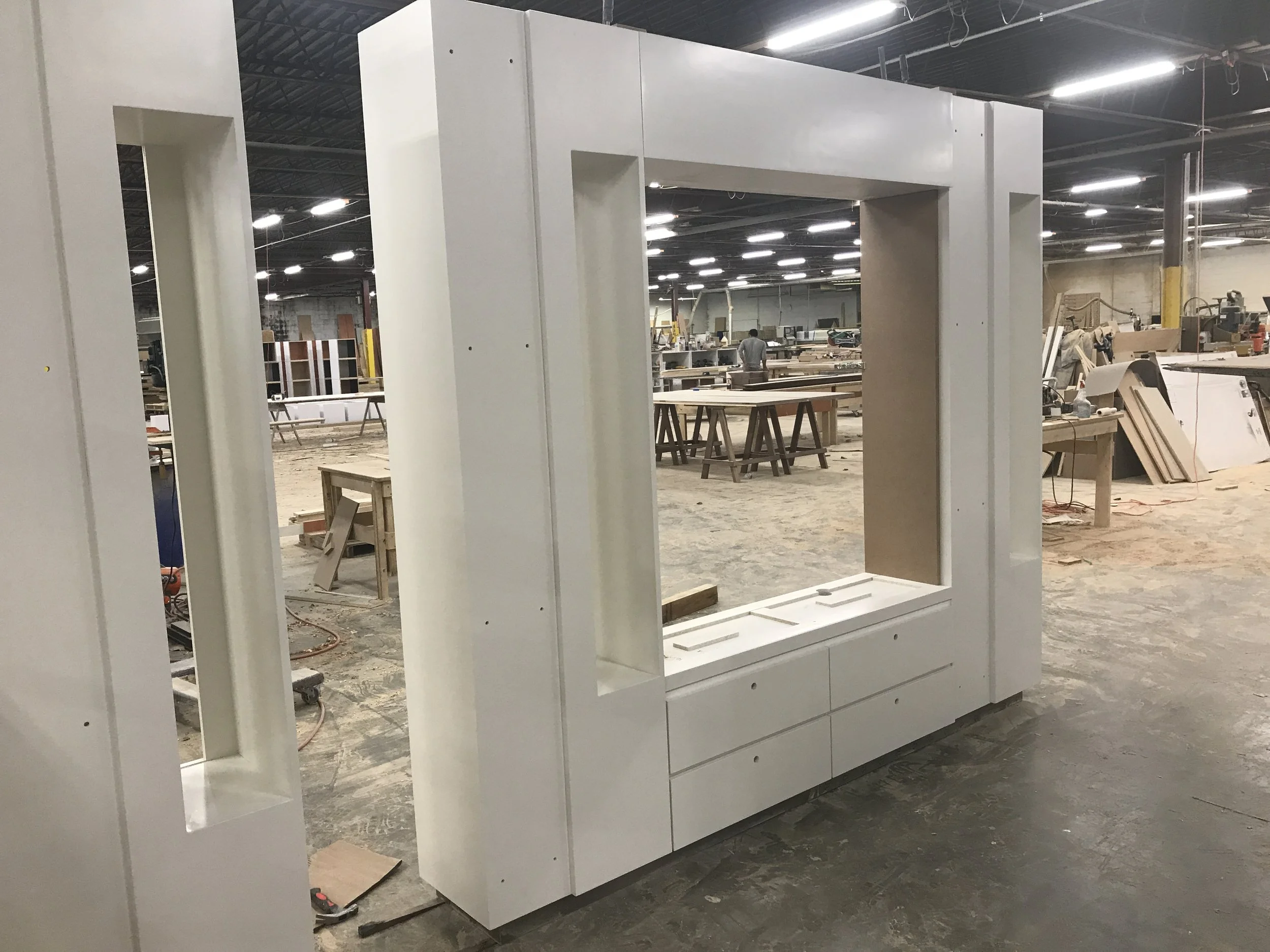 In a large industrial or warehouse space, a person works on assembling a white wardrobe or cabinet with open shelves and drawers. There are various woodworking tools and materials scattered around, with tables and equipment in the background.