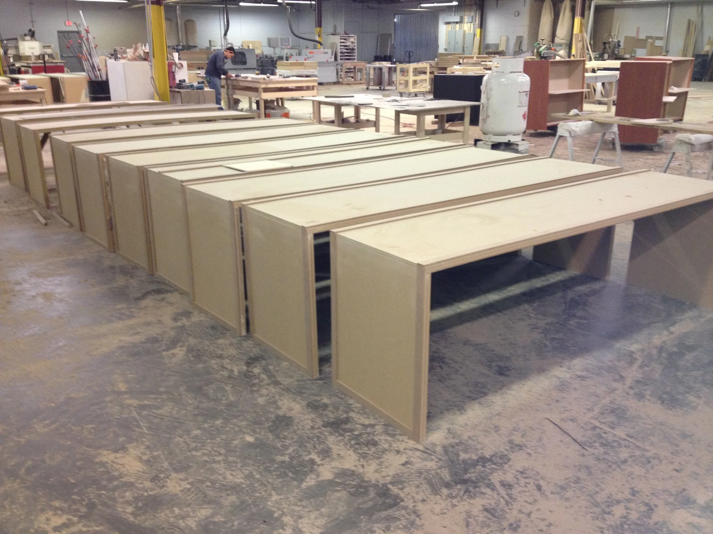 A woodworking workshop with multiple partially assembled wooden cabinets arranged on the floor and workbenches. A person is working at a workbench in the background, surrounded by tools and materials.