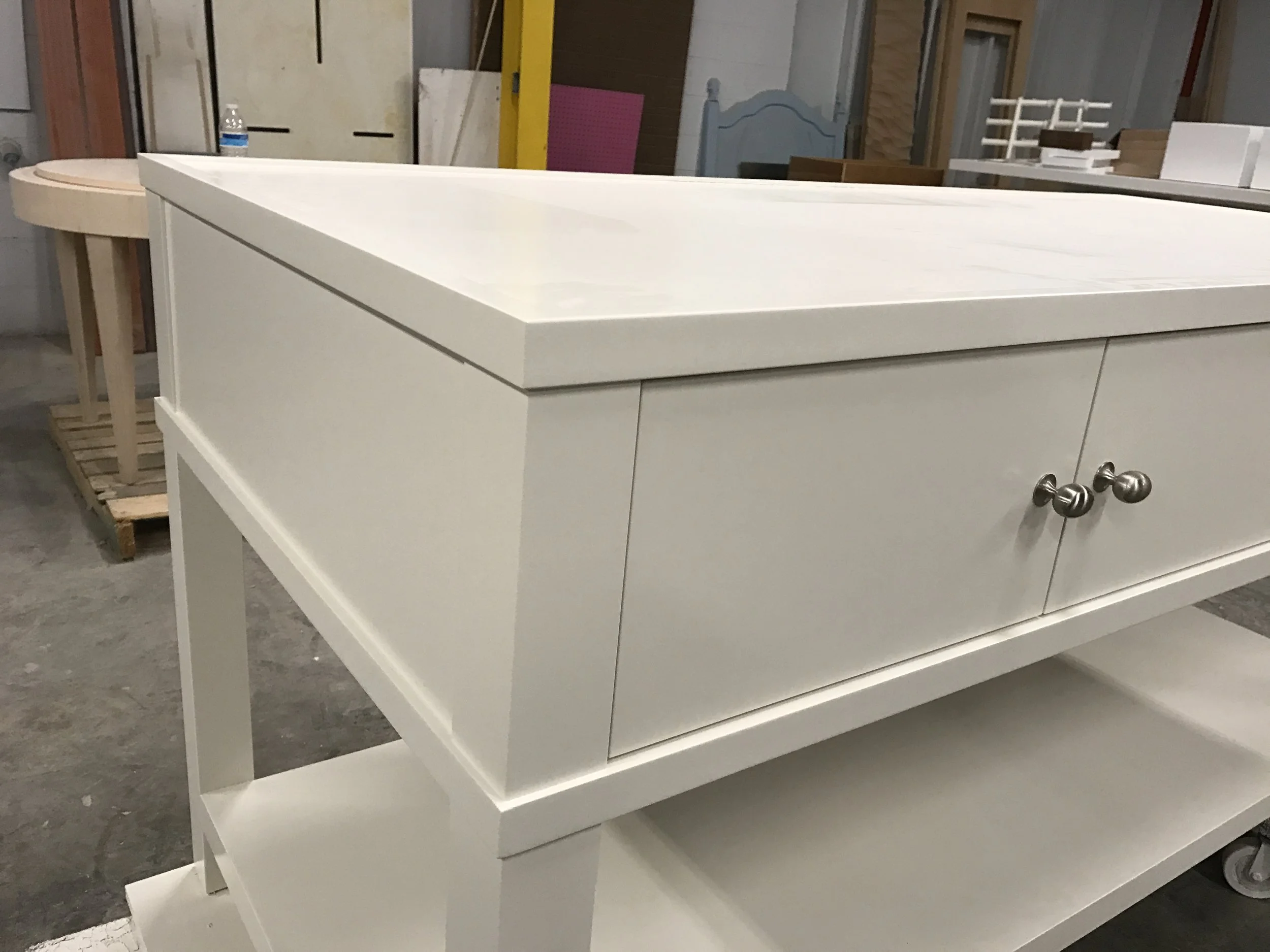 White wooden sideboard with drawers and metal knobs, placed in a workshop or store, surrounded by construction materials and other furniture.