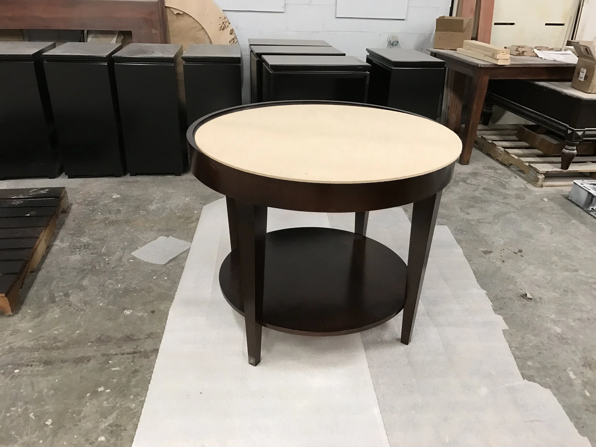 A round two-tiered table with a cream top and black legs standing on a white cloth, in a workshop with black cabinets, wooden pallets, and boxes in the background.